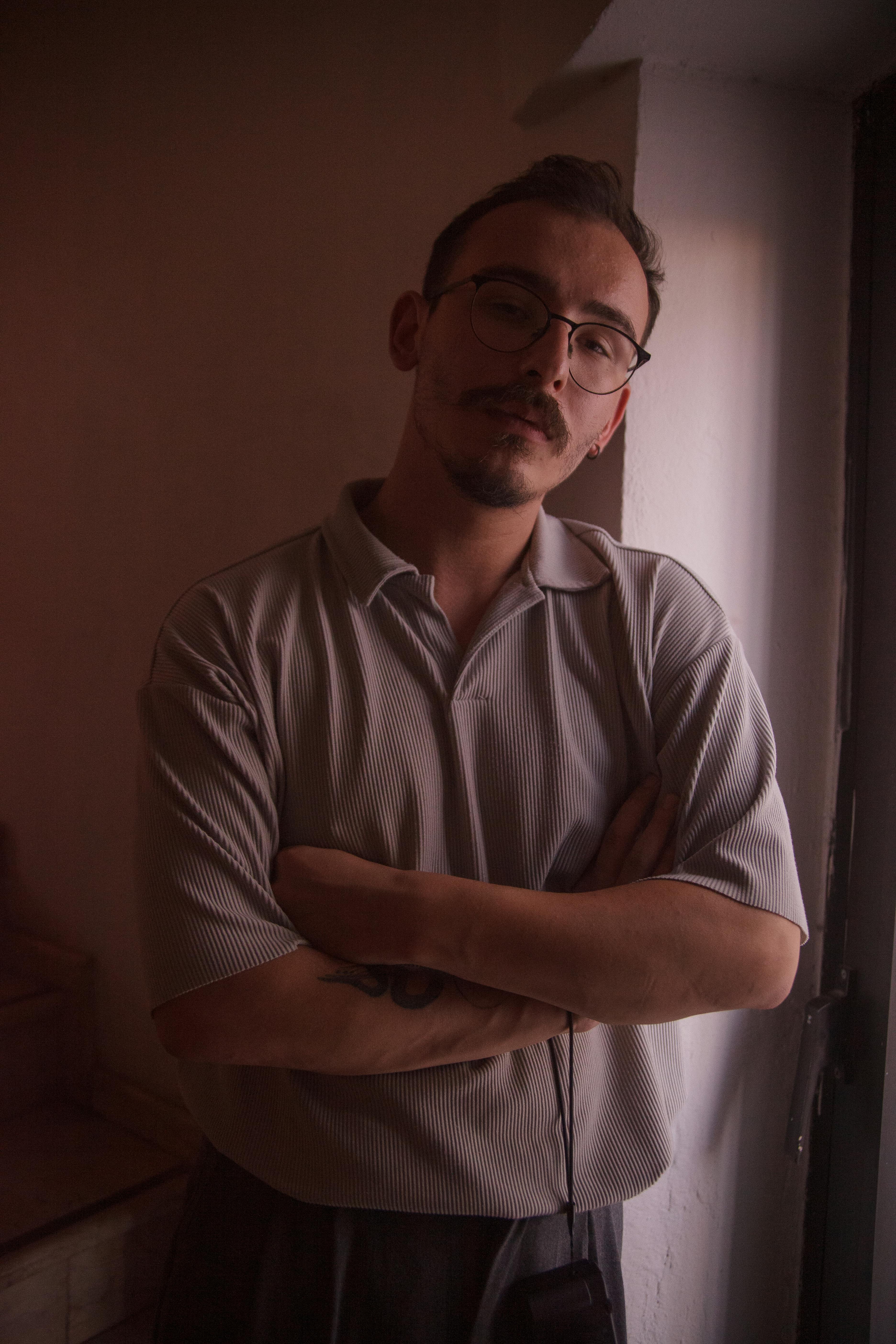 Moody indoor portrait of a man with glasses and mustache, crossing arms.