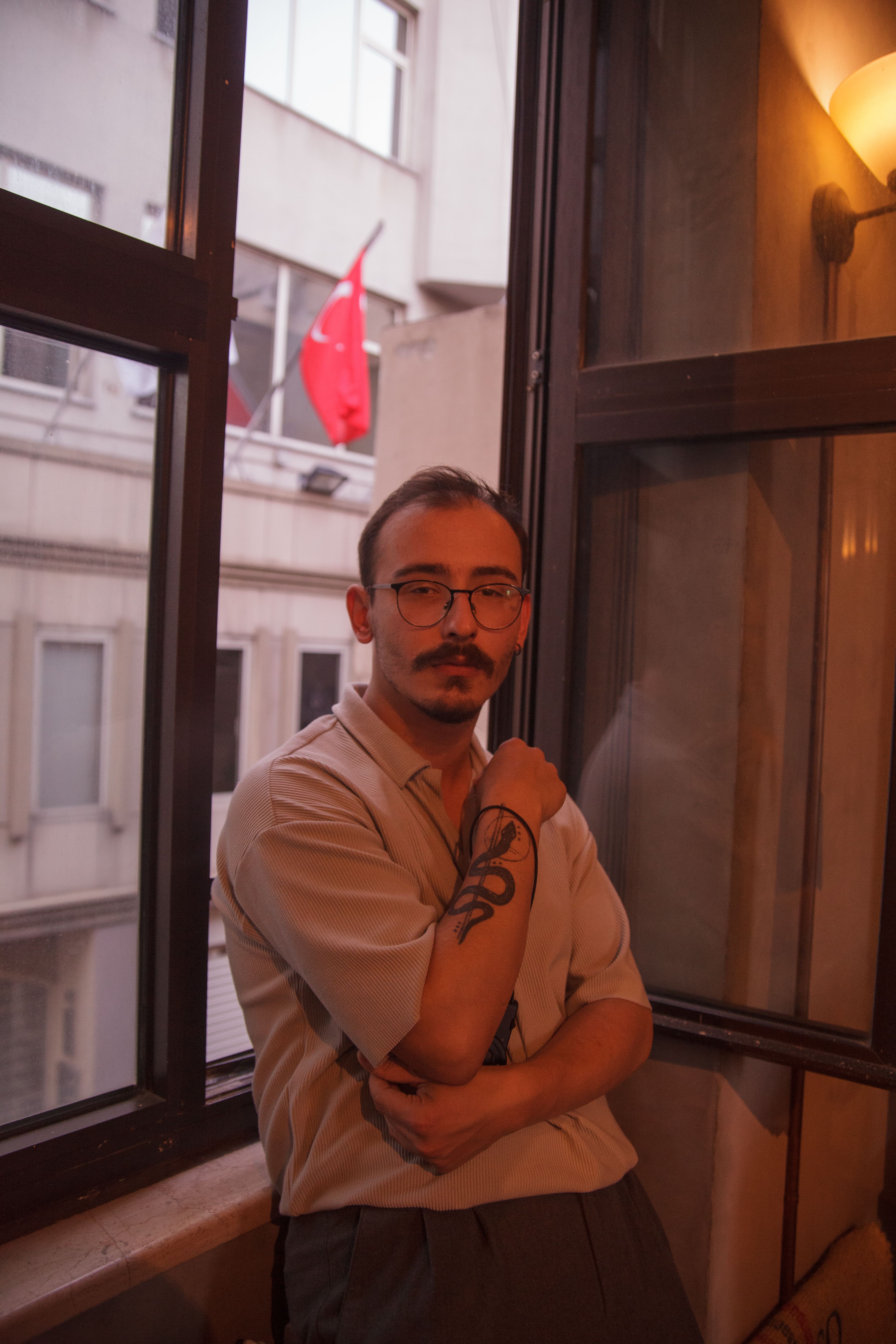 Portrait of a young man with glasses and tattoo, posing by a window in an urban setting.