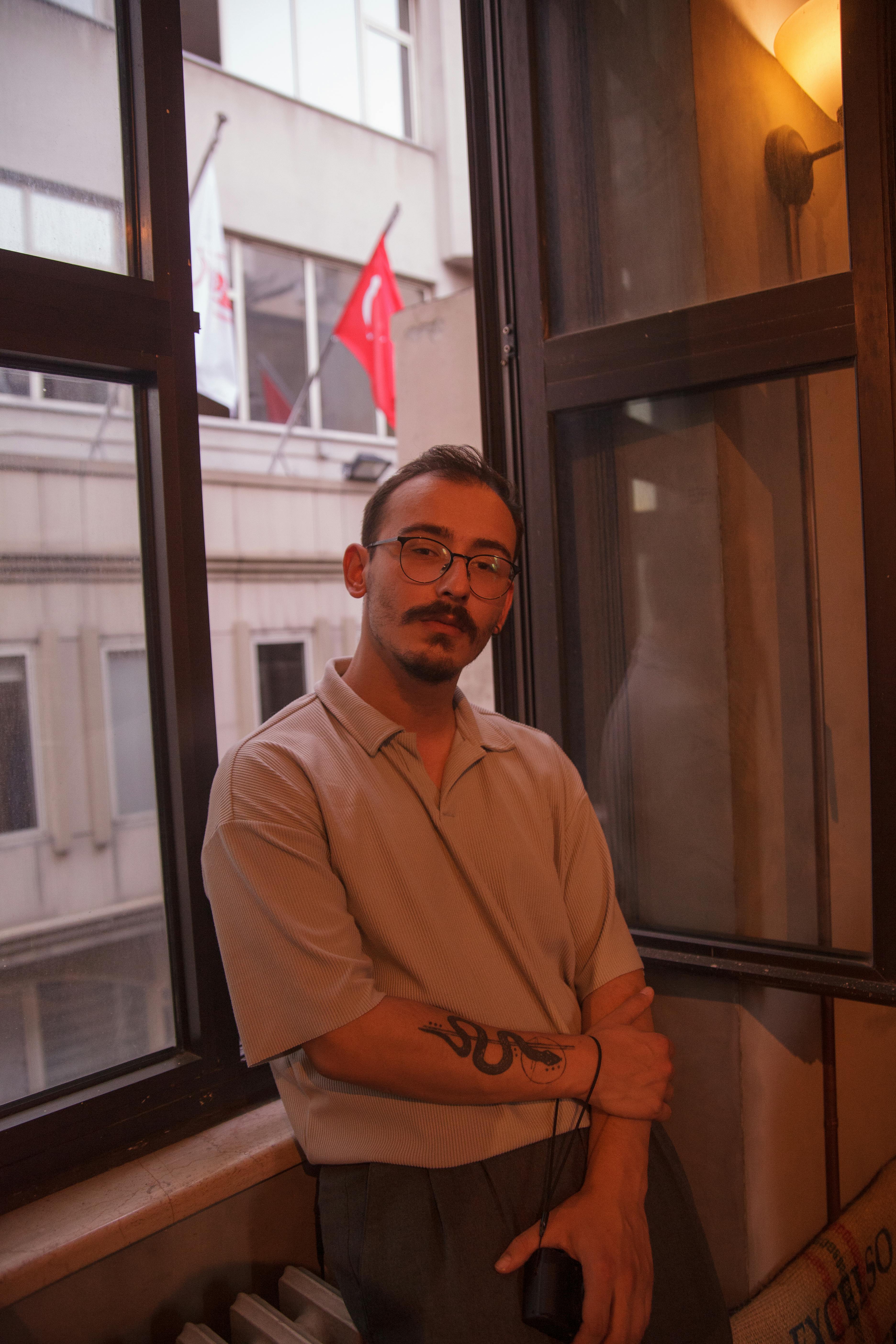 Moody portrait of a man with glasses and mustache, leaning by a city window.