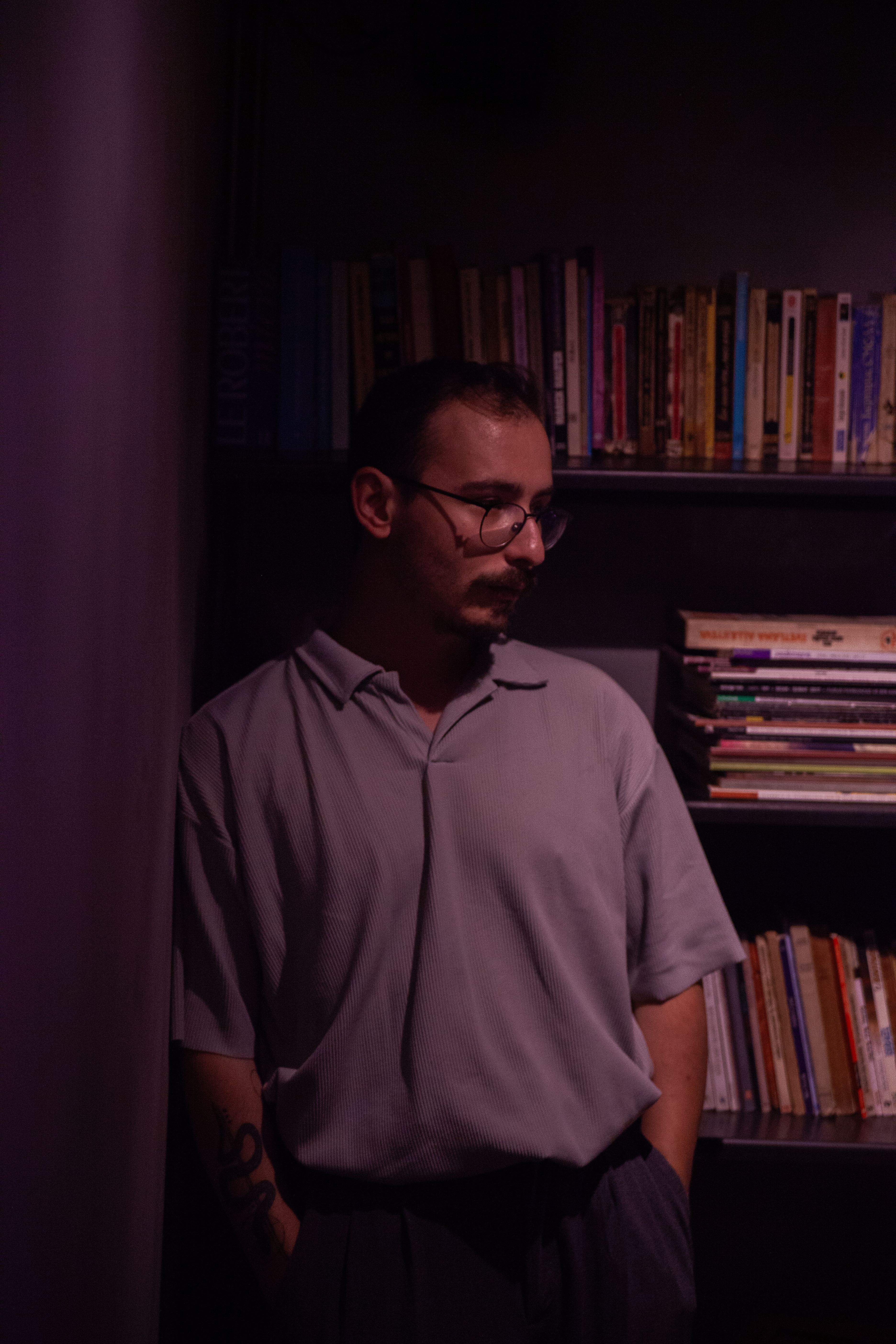 Portrait of a young man with glasses, deep in thought against a dimly lit bookshelf.