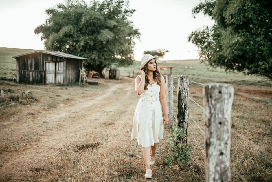 Young woman in white dress walking along a rustic path by a fence, exuding a serene and fashionable vibe.