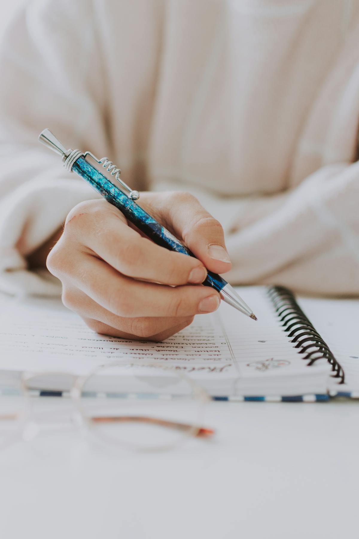 picture taken of a hand holding a pen in process of writing on a paper. the person is wearing a white cardigan and the pen is blue.