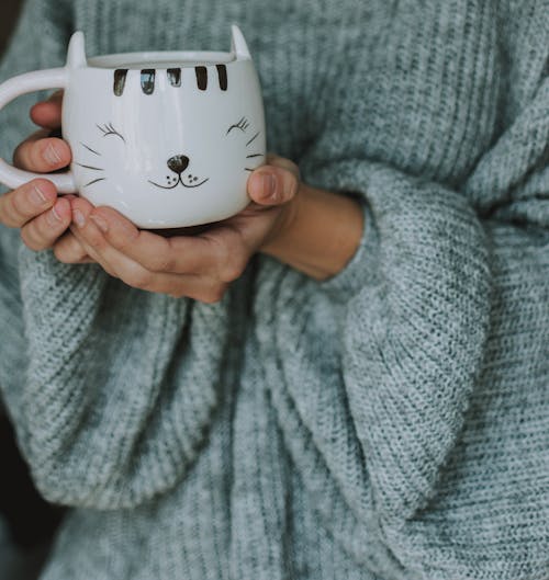 Unrecognizable female in warm long sleeved knit grey sweater with cup of hot coffee at home