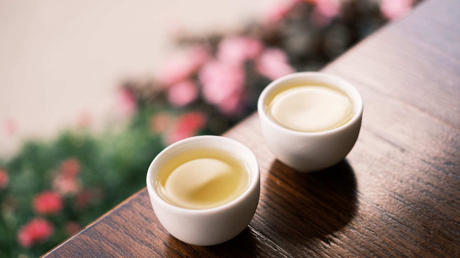 Two white cups of tea on a wooden table with flowers in the background