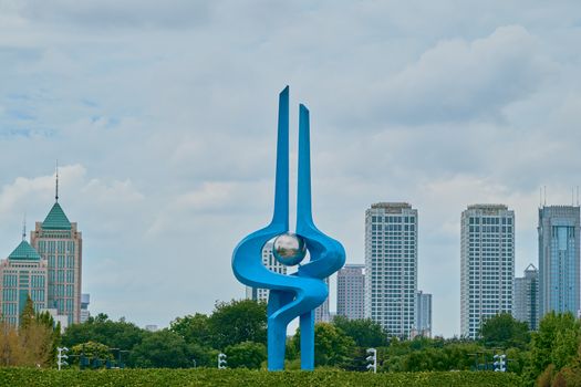 Blue modern sculpture set against a city skyline under a cloudy sky.