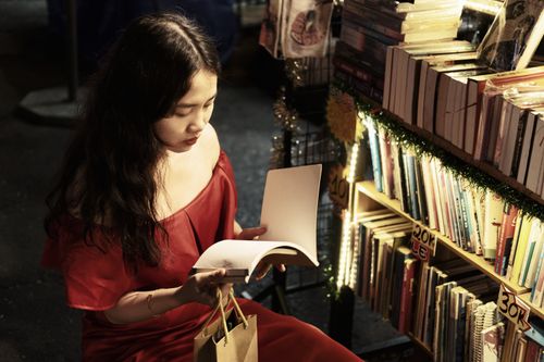 Free Woman in red dress reading a book at a dimly lit night market bookstall, surrounded by books. Stock Photo