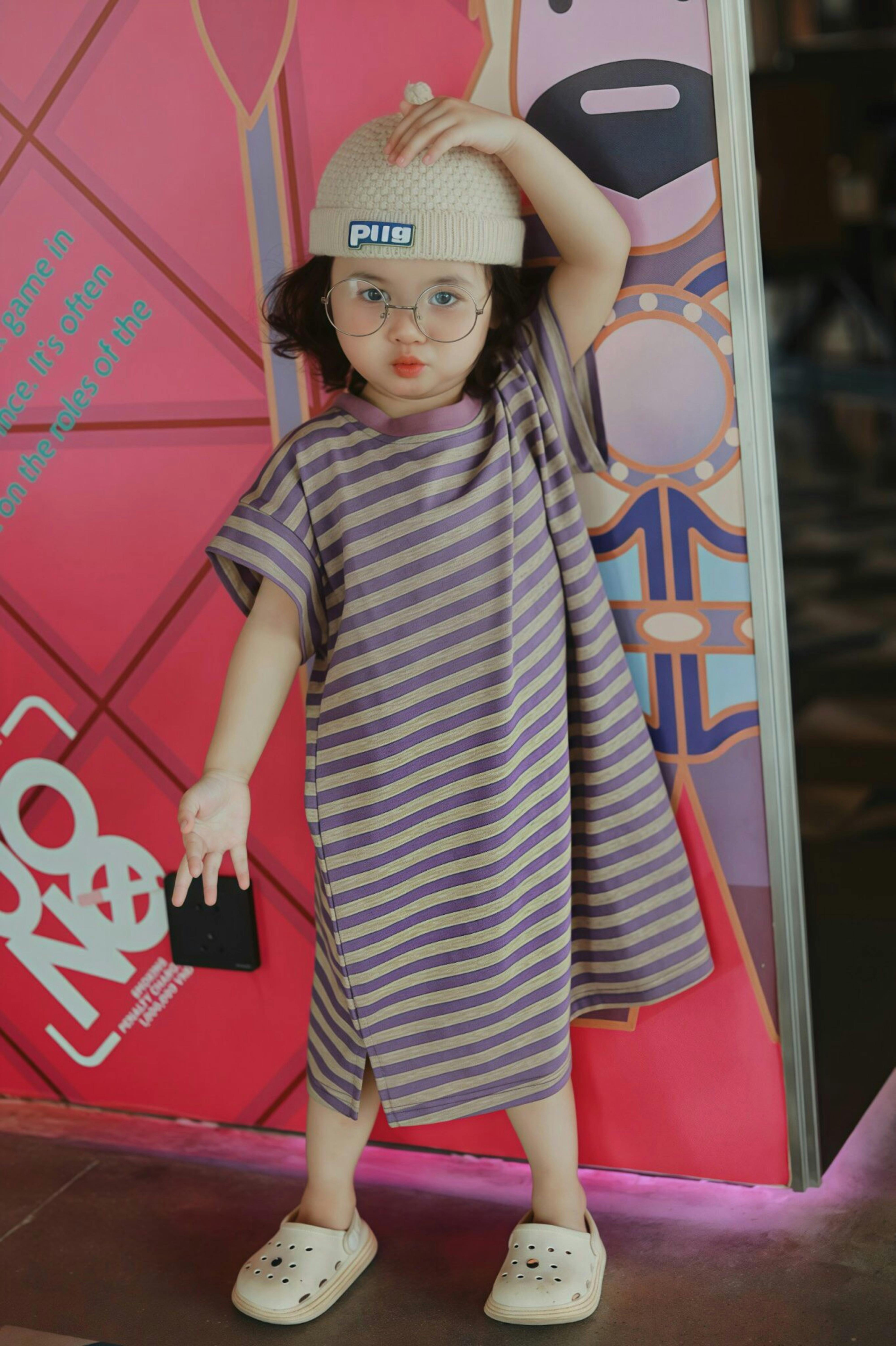 Free Cute child wearing a striped dress and glasses indoors, posing playfully against a colorful backdrop. Stock Photo