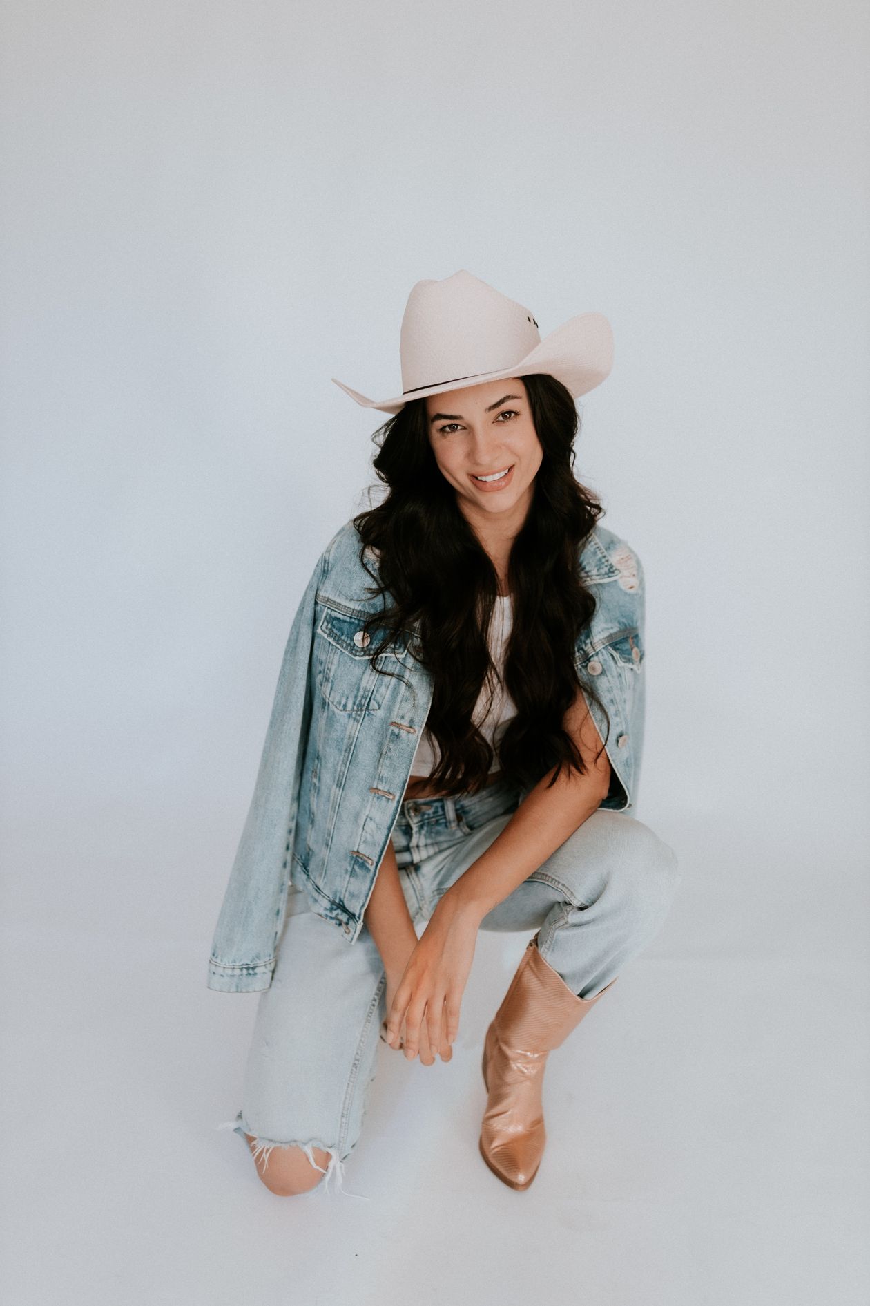 Smiling woman in a light-wash denim jacket, cream cowboy hat, and tan western boots