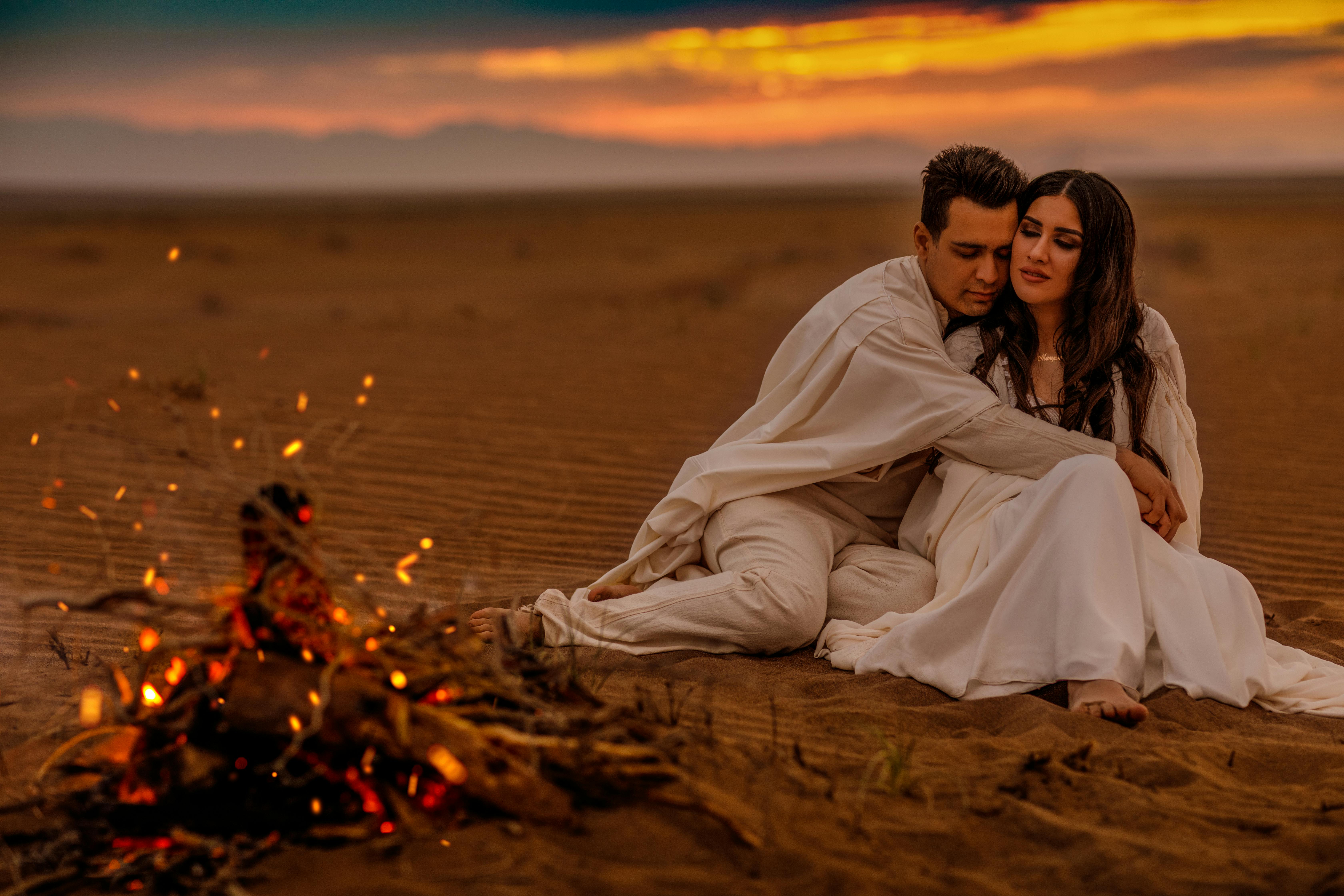 A romantic couple enjoying a desert sunset with a warm bonfire.