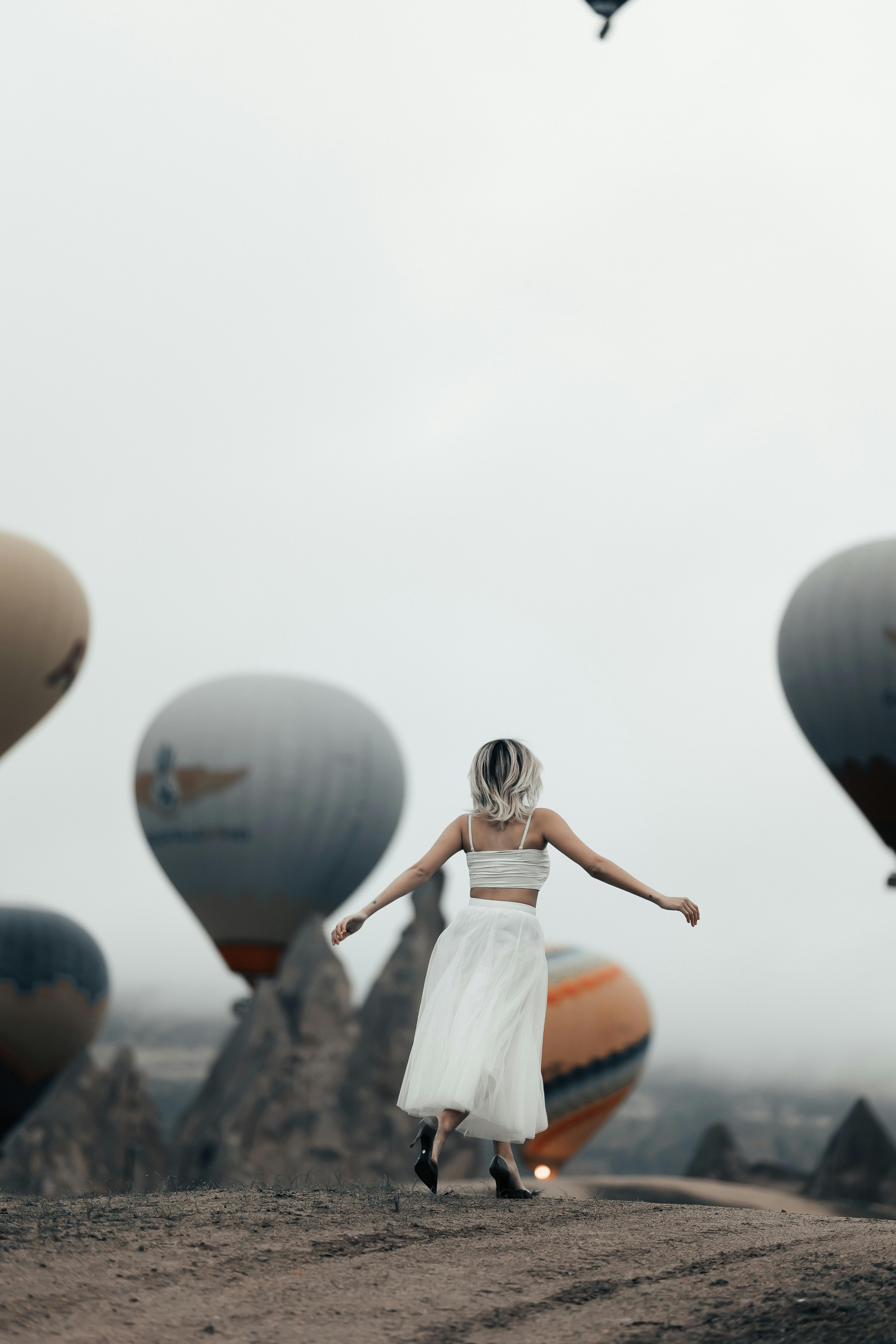 A woman in a white dress stands joyfully among Cappadocia's iconic hot air balloons.