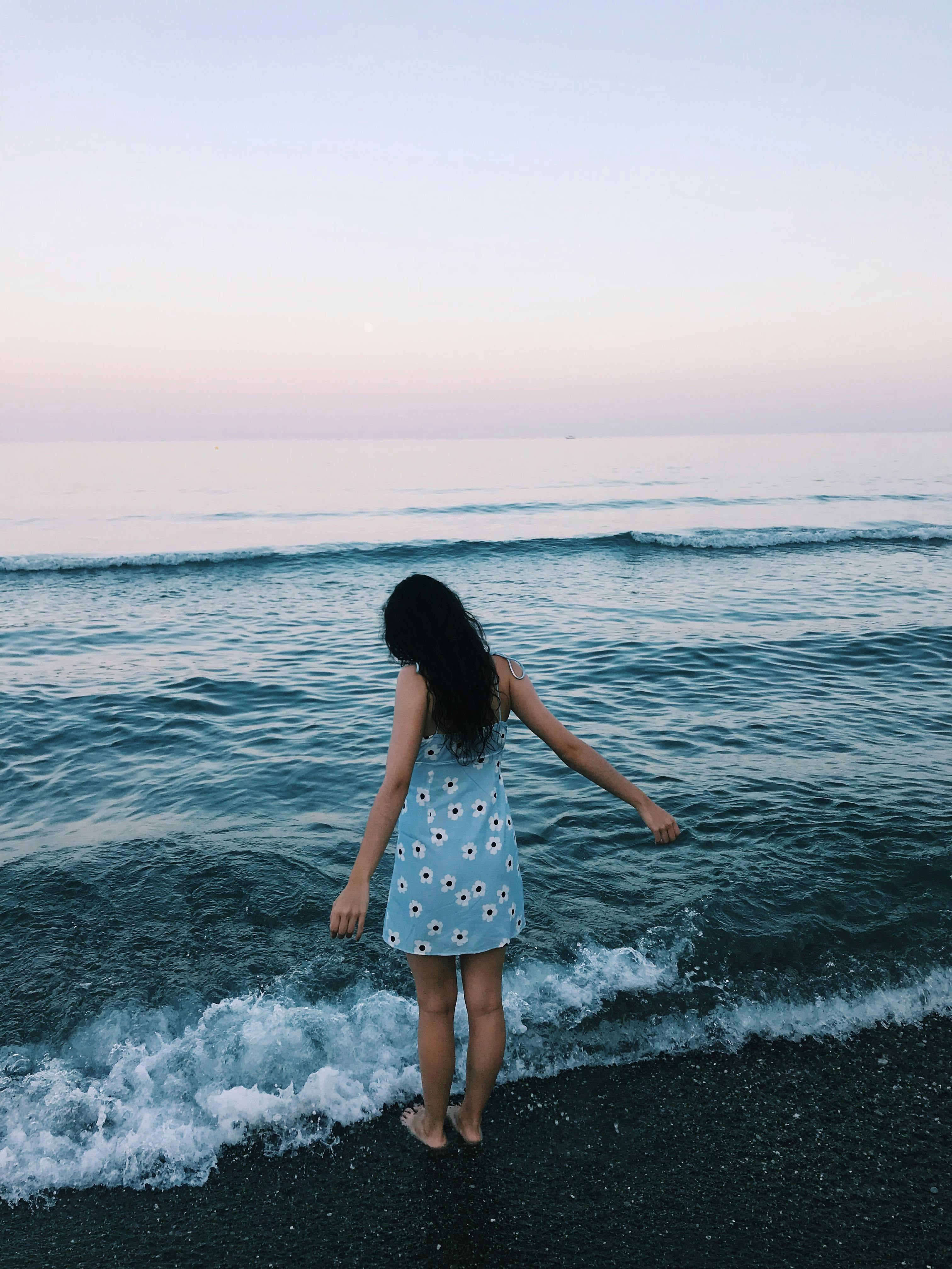 A woman stands by the ocean in a floral dress, embracing the tranquility of sunset.