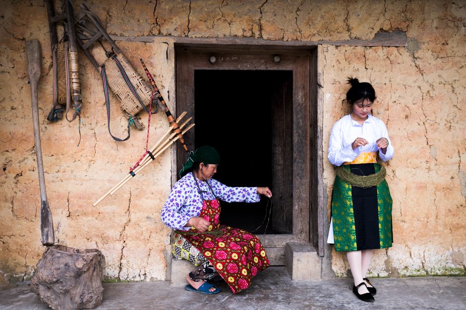 Two women in traditional clothing sit and stand at a village entrance.
