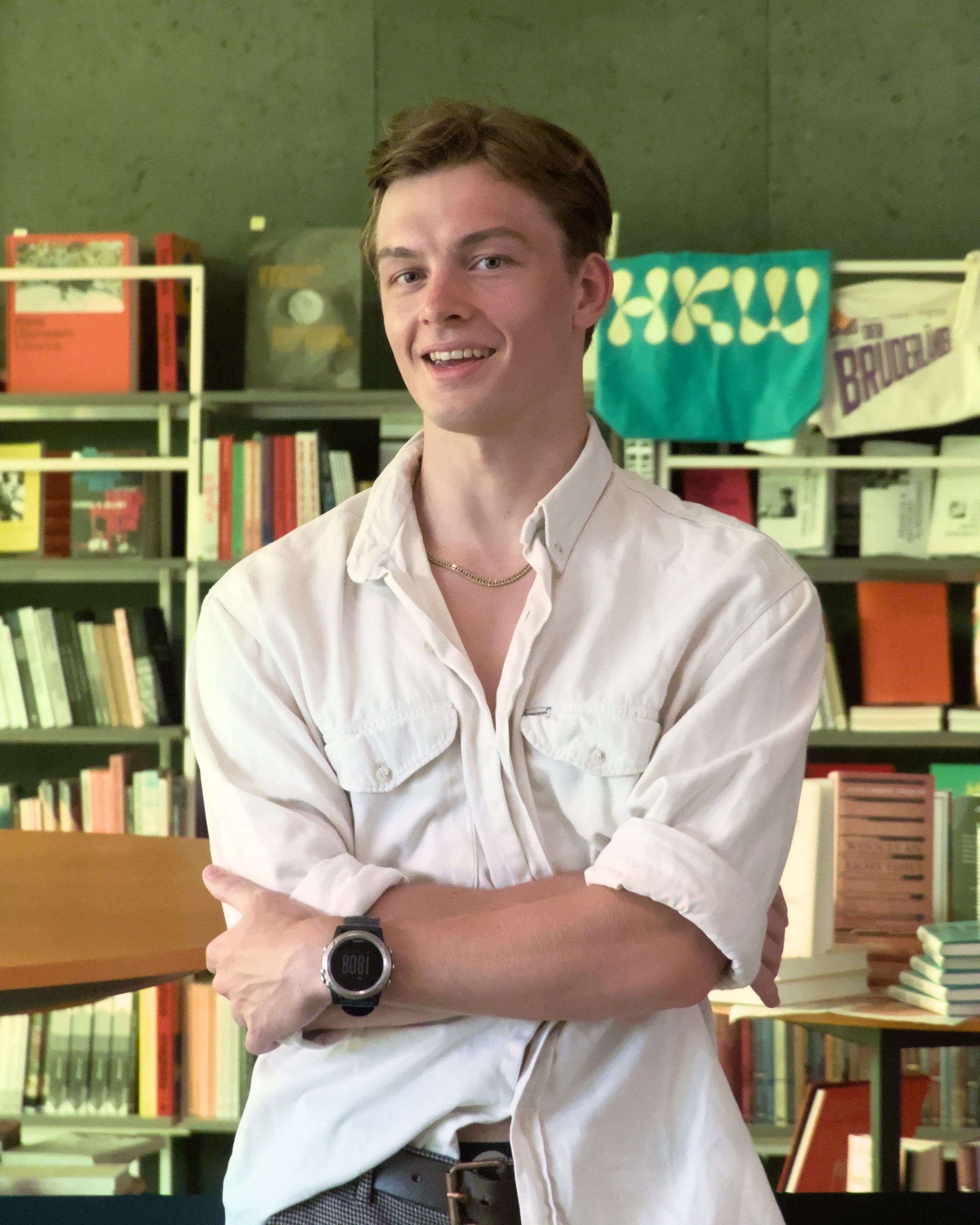 Portrait of a young man standing in a Berlin library, smiling with folded arms.