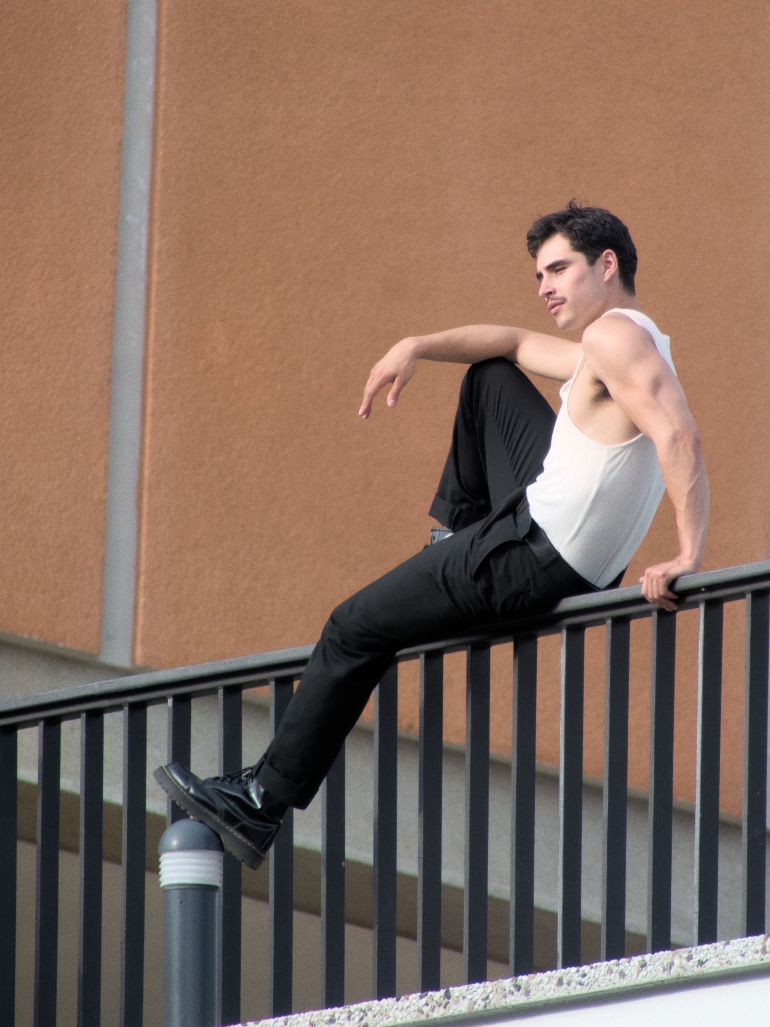 A fashionable man sitting on a railing outdoors in Berlin, showcasing confidence.