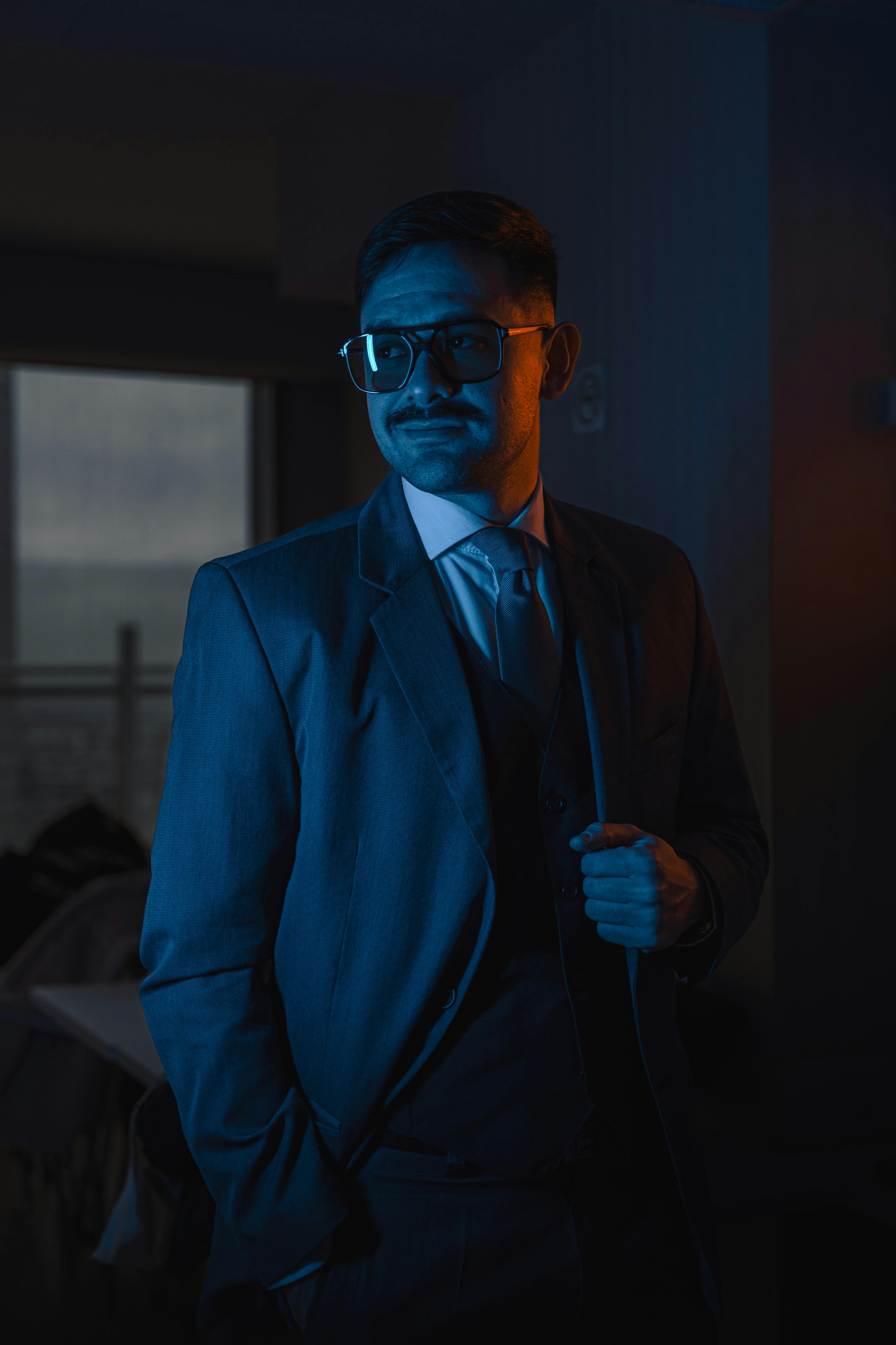 Portrait of a man with glasses in a suit, indoors with dramatic lighting, conveying sophistication.