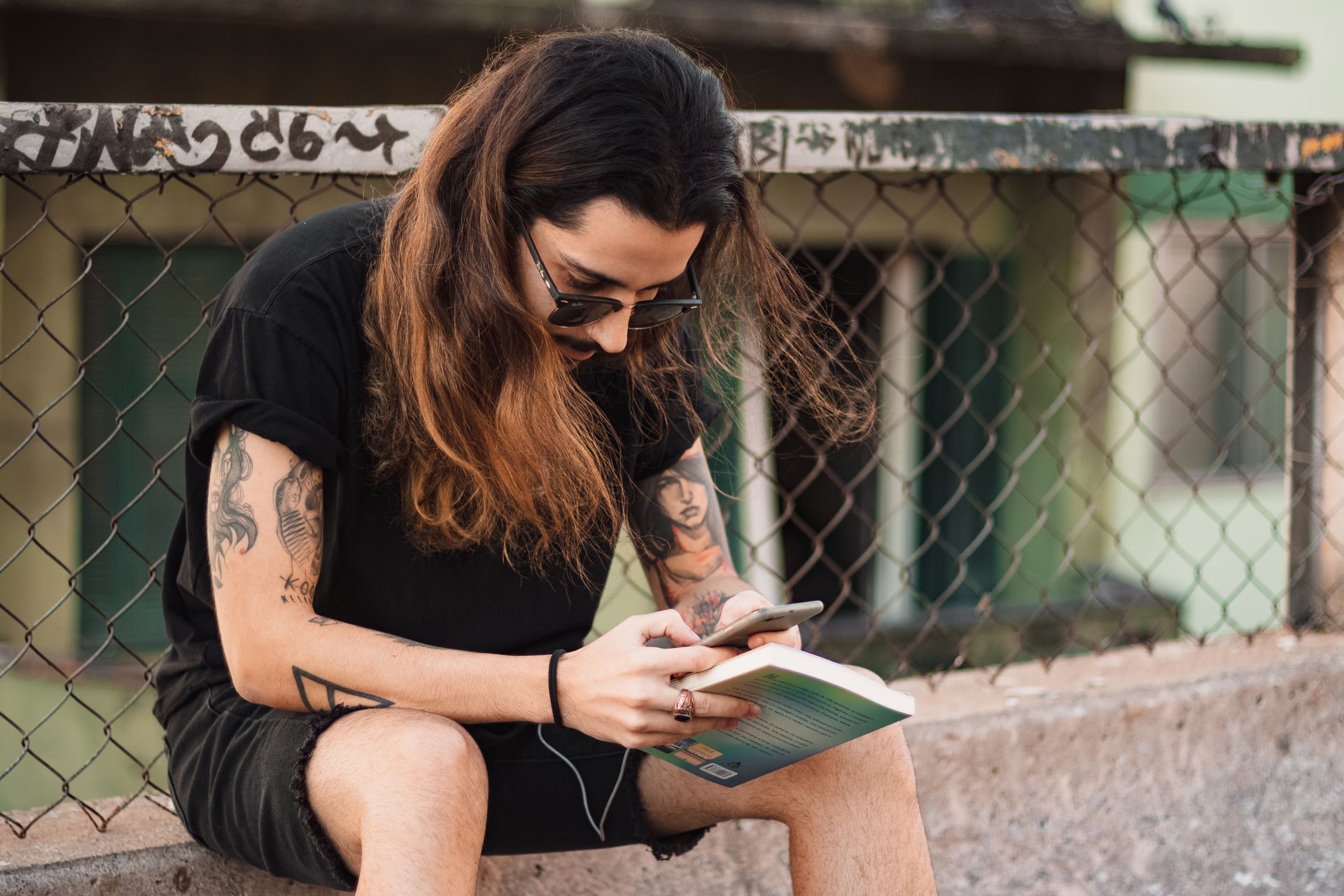 Tattooed man with long hair texting on a smartphone while sitting outdoors.