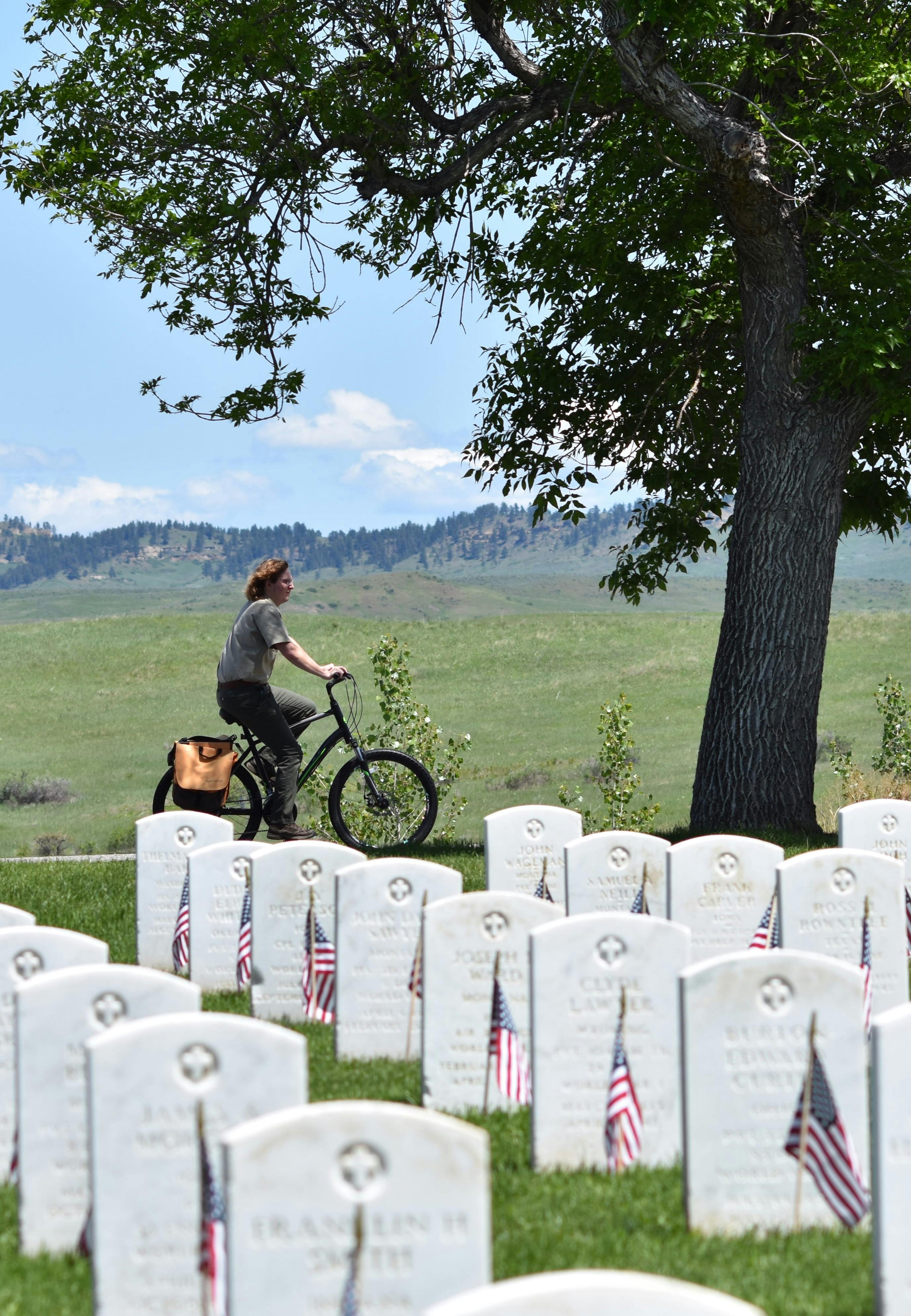 A female park ranger rides a bike with saddlebag among American-flagged headstones.