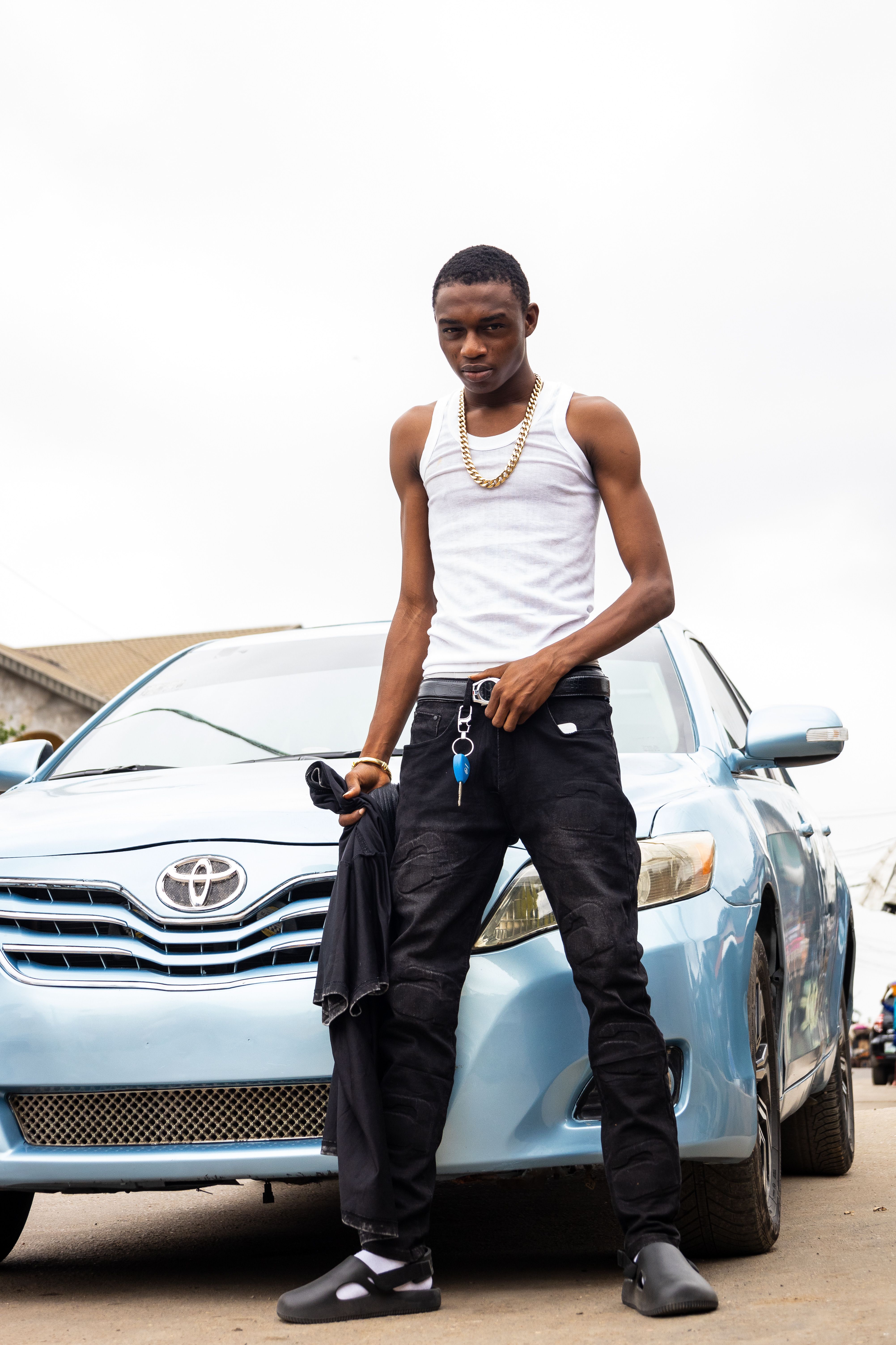 A stylish young man standing in front of a blue car wearing casual attire and accessories.