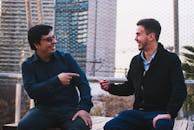 两名男子坐在户外，微笑着互相指着对方，做出友好的手势