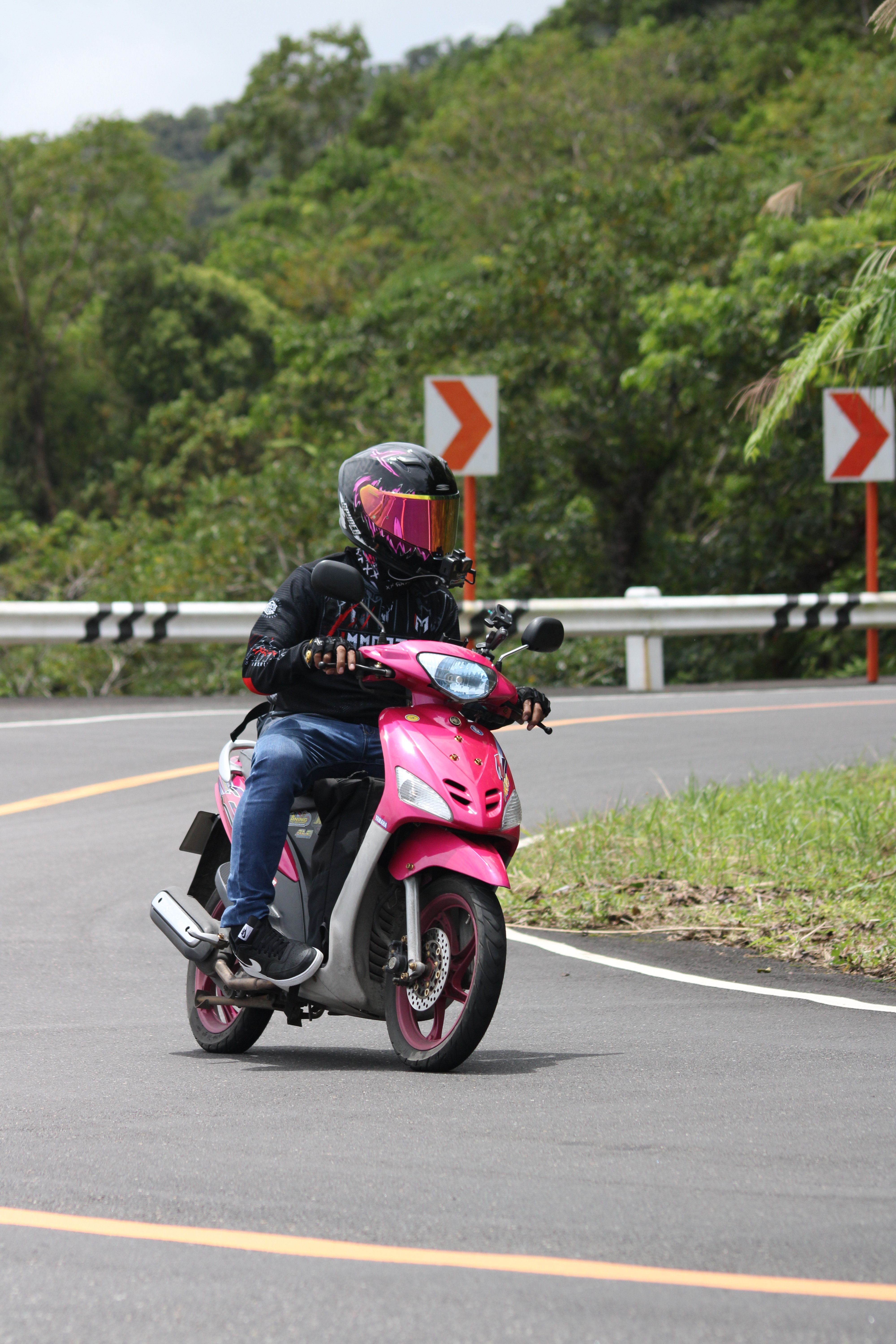 Motorcyclist in gear riding a vibrant pink scooter on a curvy, scenic road during the day.