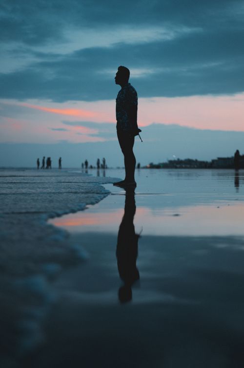 Free A lone figure stands on a beach at sunset, creating a peaceful silhouette reflection. Stock Photo