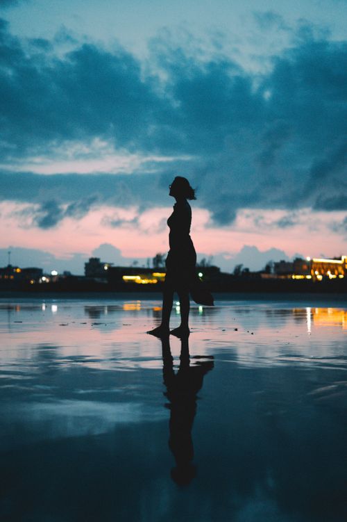 Free A serene silhouette of a woman by the water reflecting a dramatic sunset sky. Stock Photo