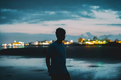 Free Silhouette of a person standing on a beach at dusk with city lights in the background. Stock Photo