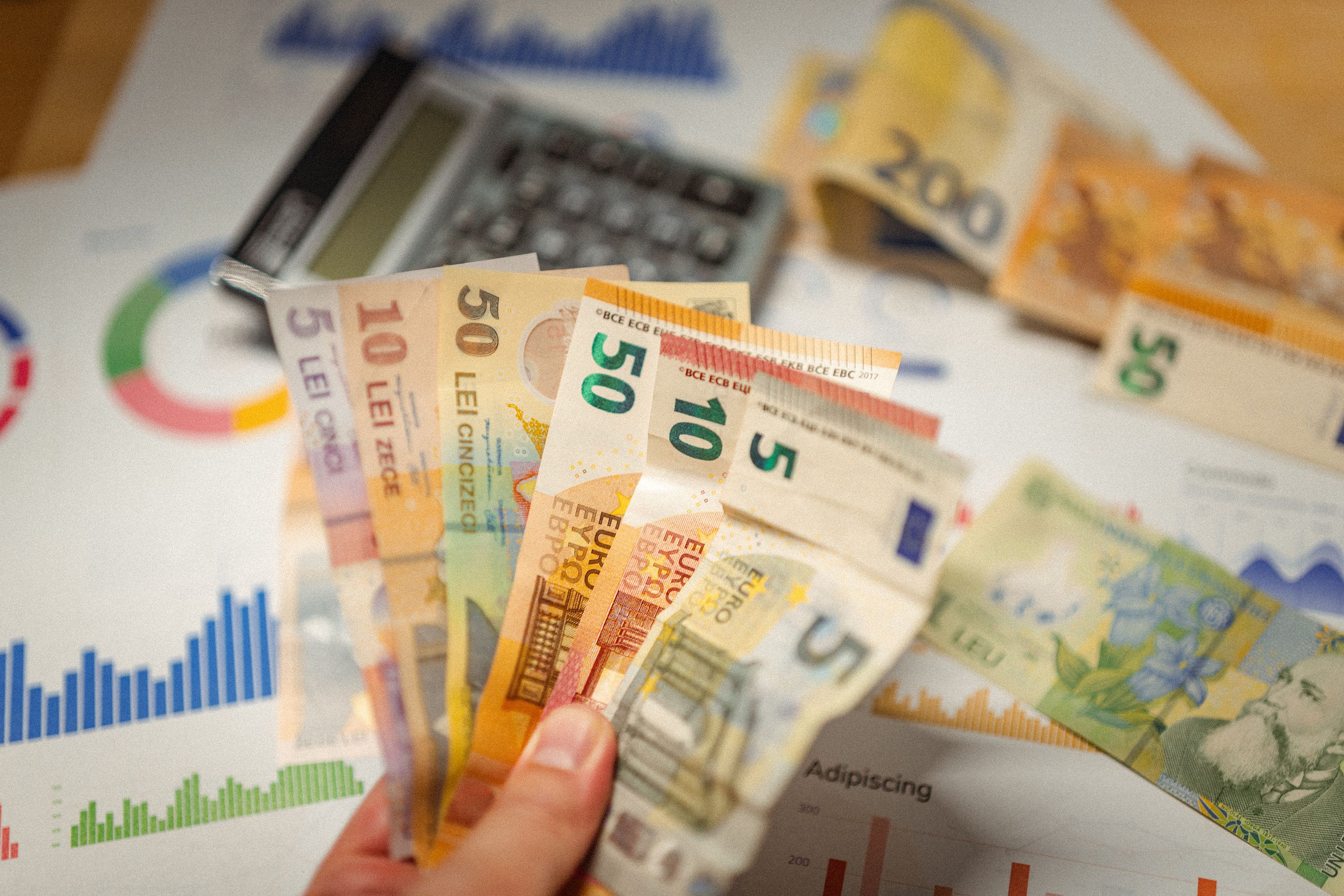 Hand holding euro banknotes over financial charts, with a calculator and other currencies in the background