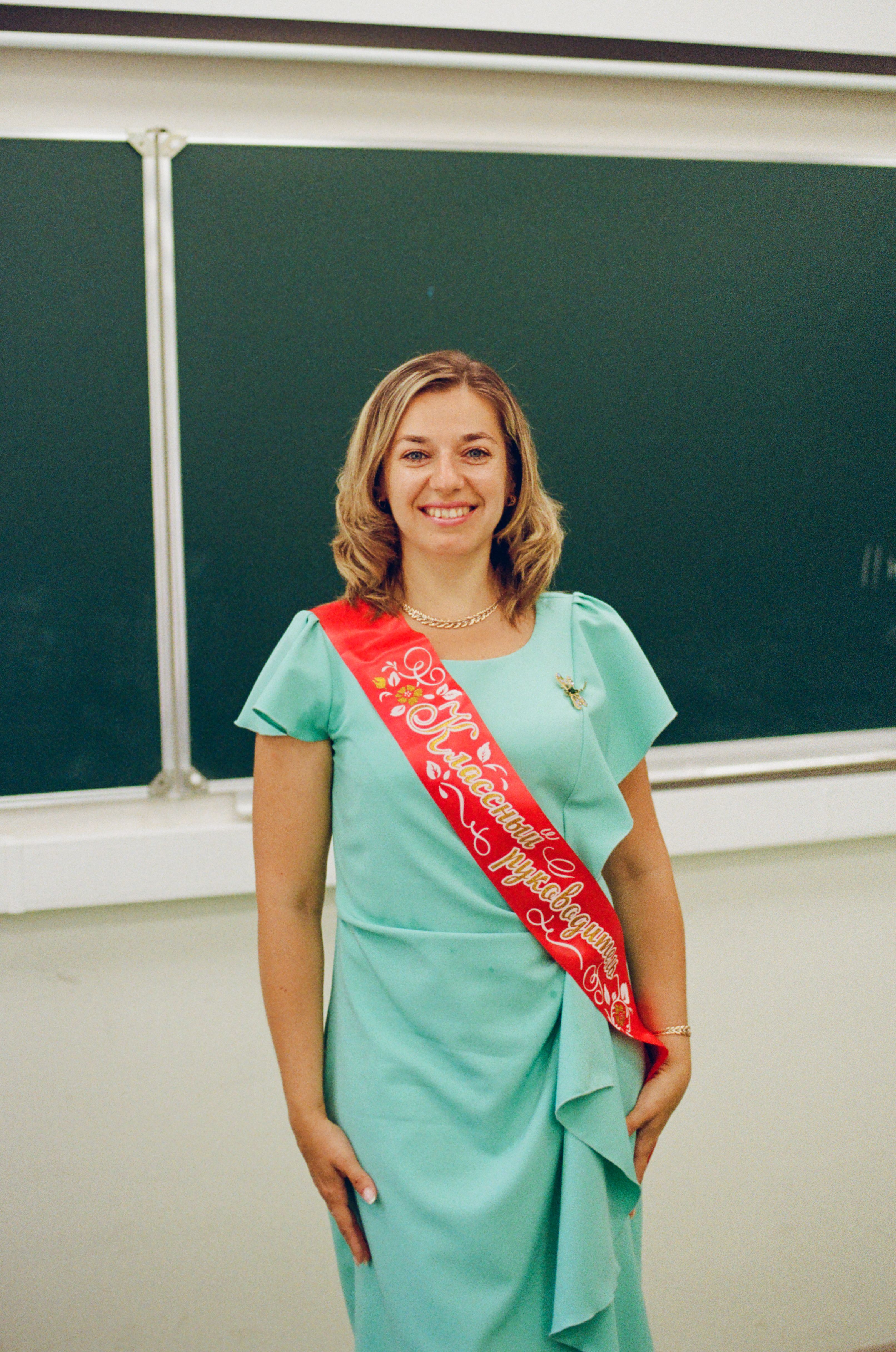 Smiling woman in a teal dress with a red sash standing in front of a chalkboard, conveying happiness.