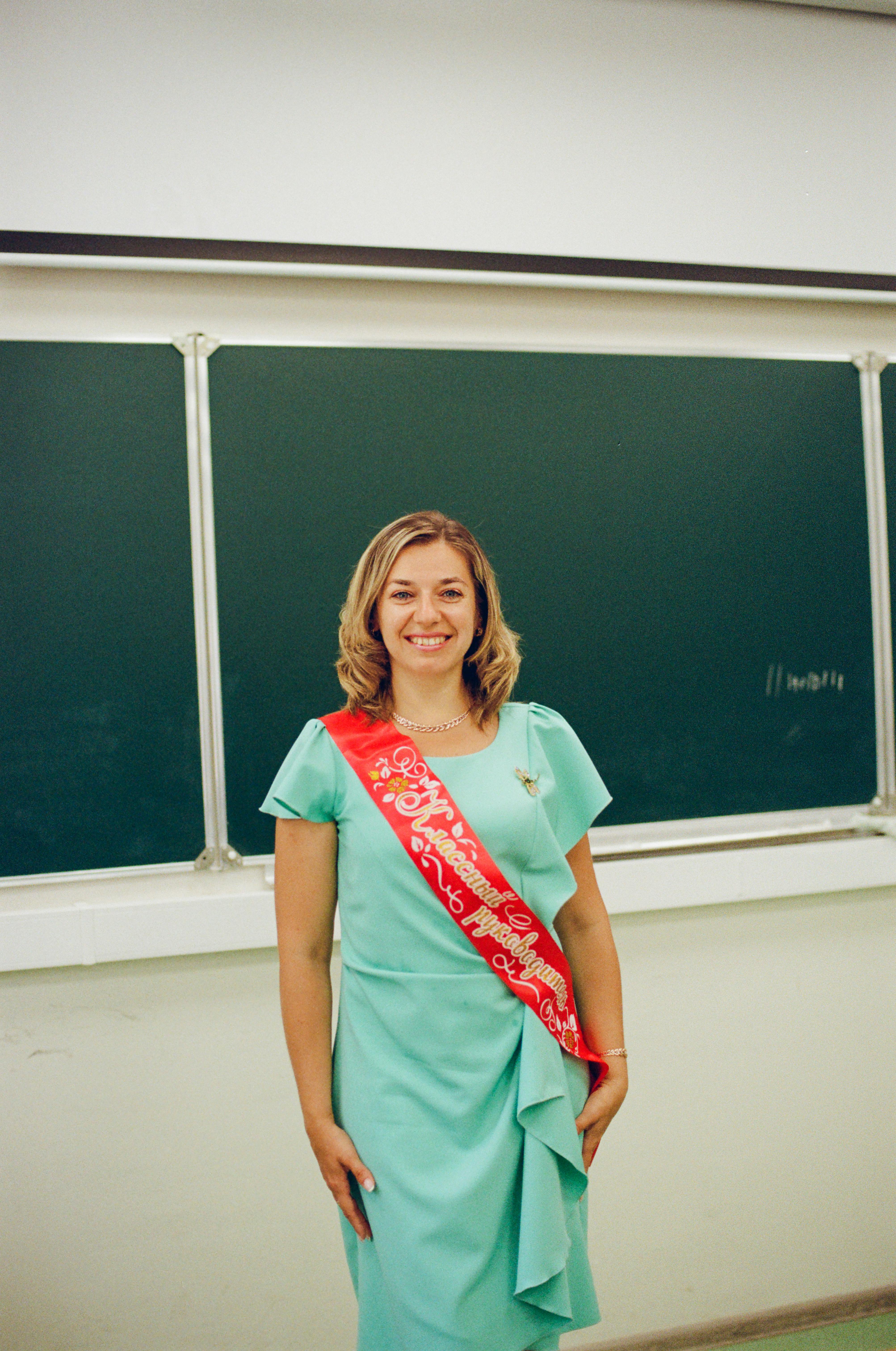 A smiling woman in a mint dress with a colorful sash stands in front of a classroom chalkboard.
