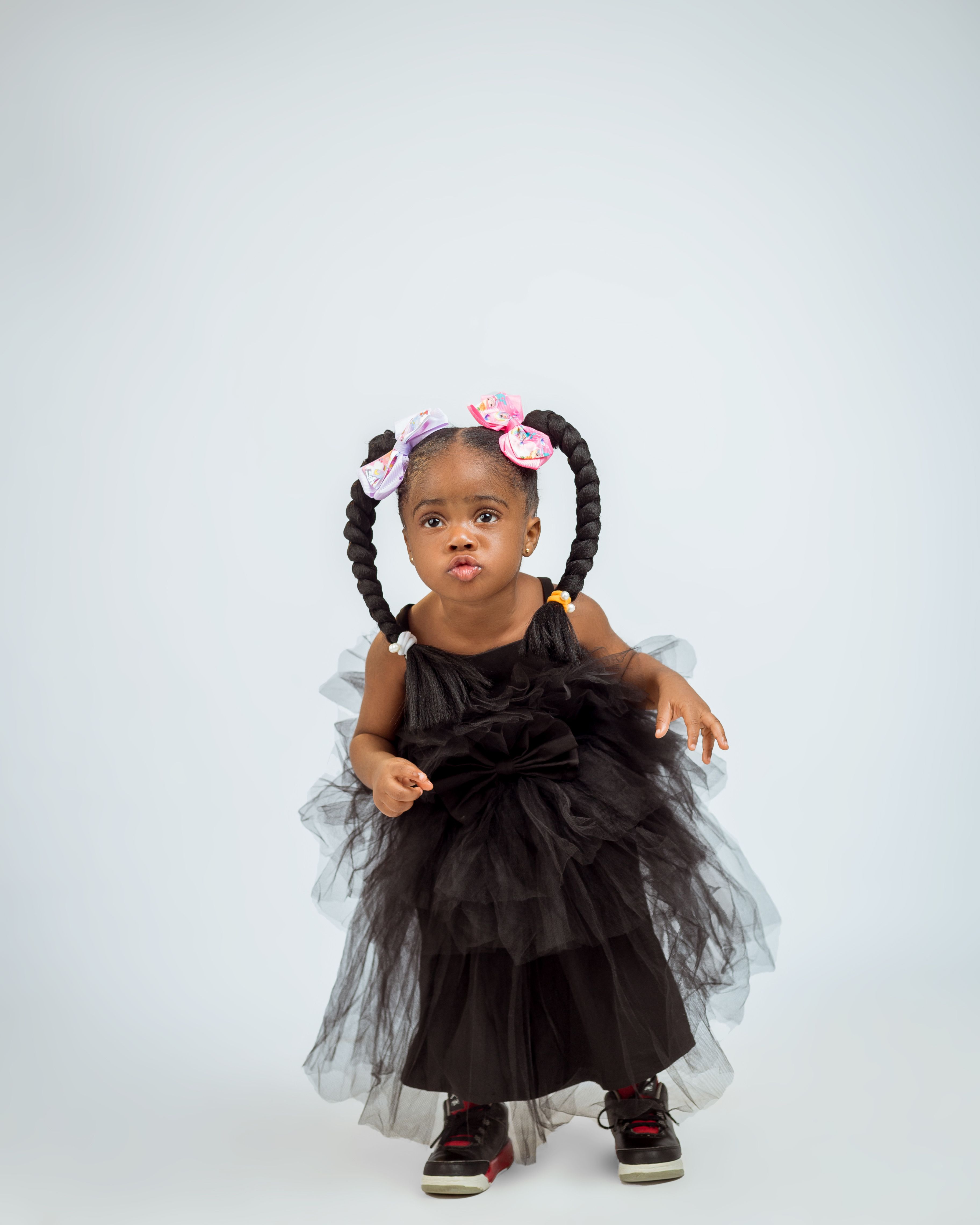 Cute toddler in a stylish black tulle dress and colorful braids posing playfully.