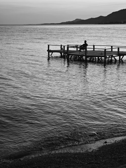 Free A calming black and white image of a person sitting on a pier at dawn, reflecting in water. Stock Photo