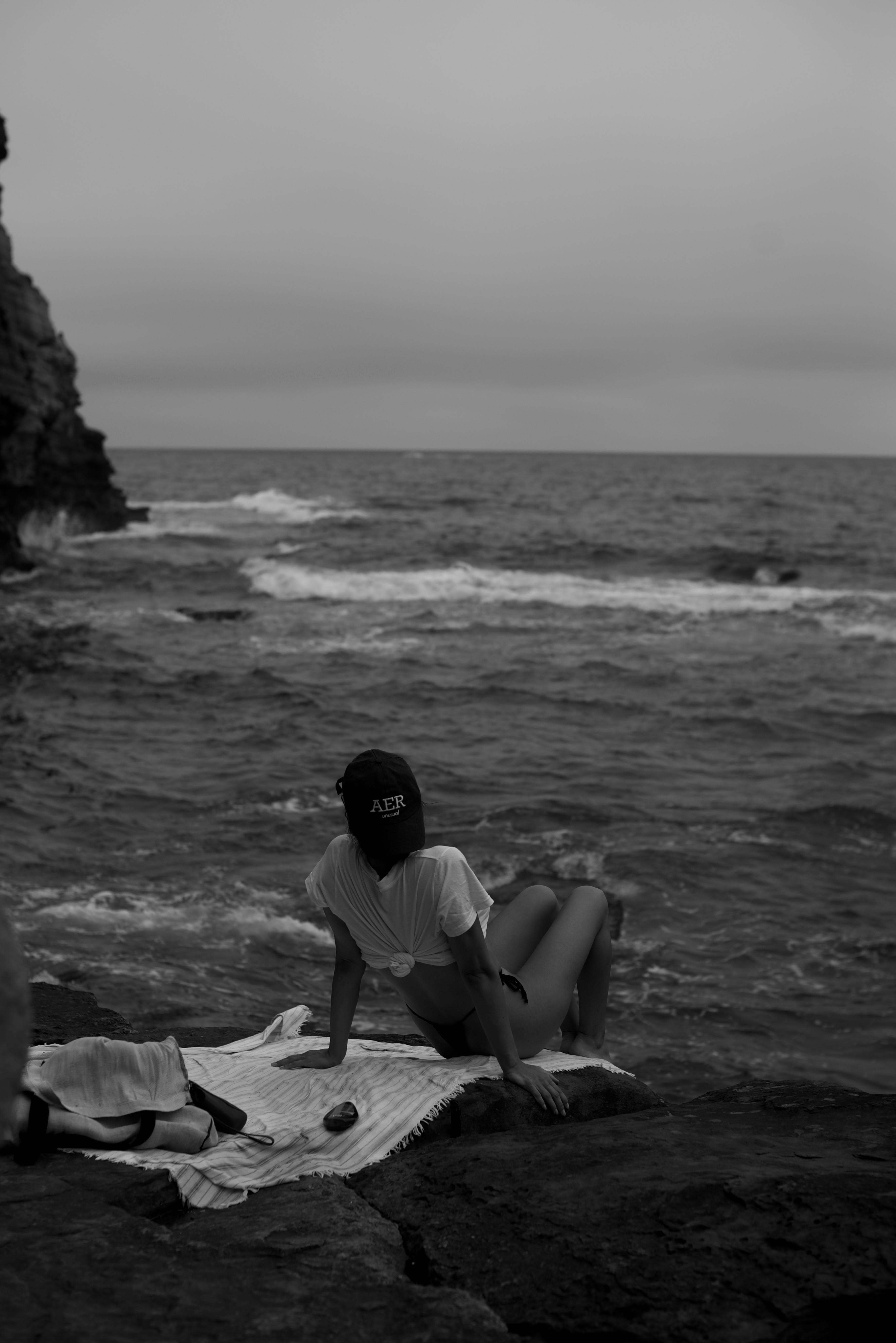 A woman relaxes by the ocean on a rocky seashore at sunset, capturing serenity and nature.