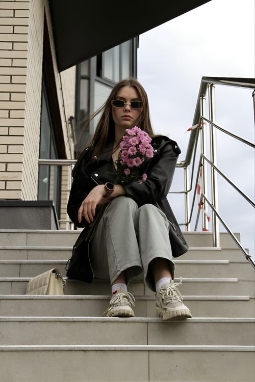 Free Fashionable woman in a leather jacket holding flowers, sitting on stairs in an urban setting. Stock Photo