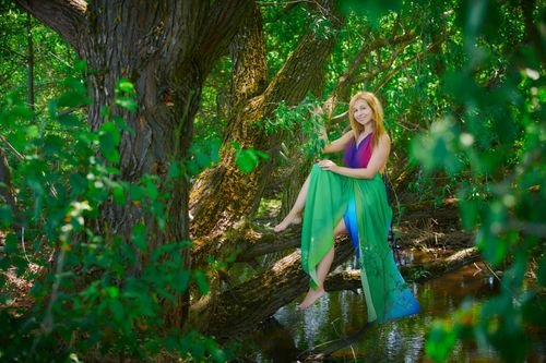 Free A woman in a colorful dress smiles while sitting on a tree in a lush forest. Stock Photo