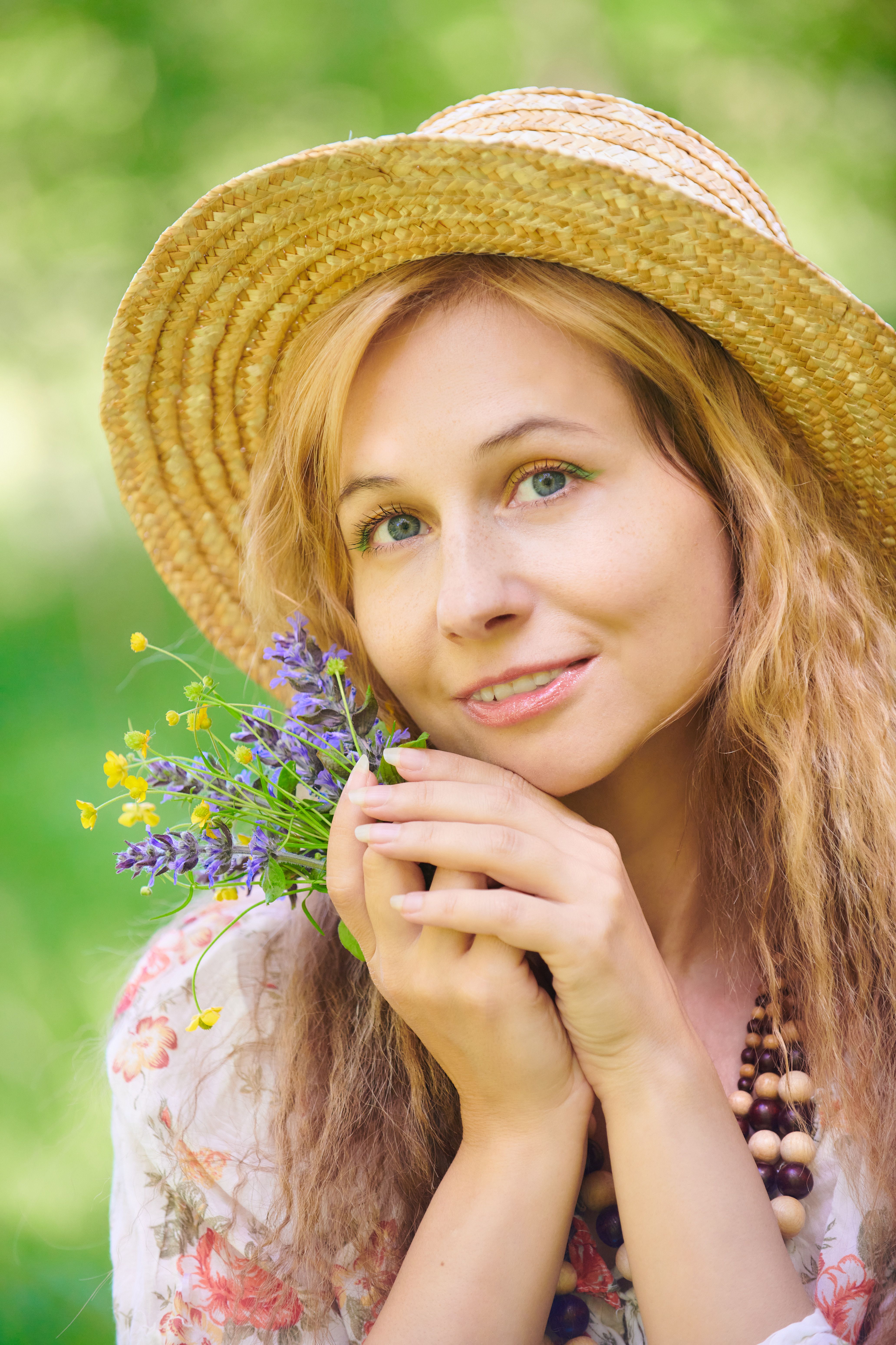 Young woman in a straw hat holding a bouquet of wildflowers, smiling amidst greenery.