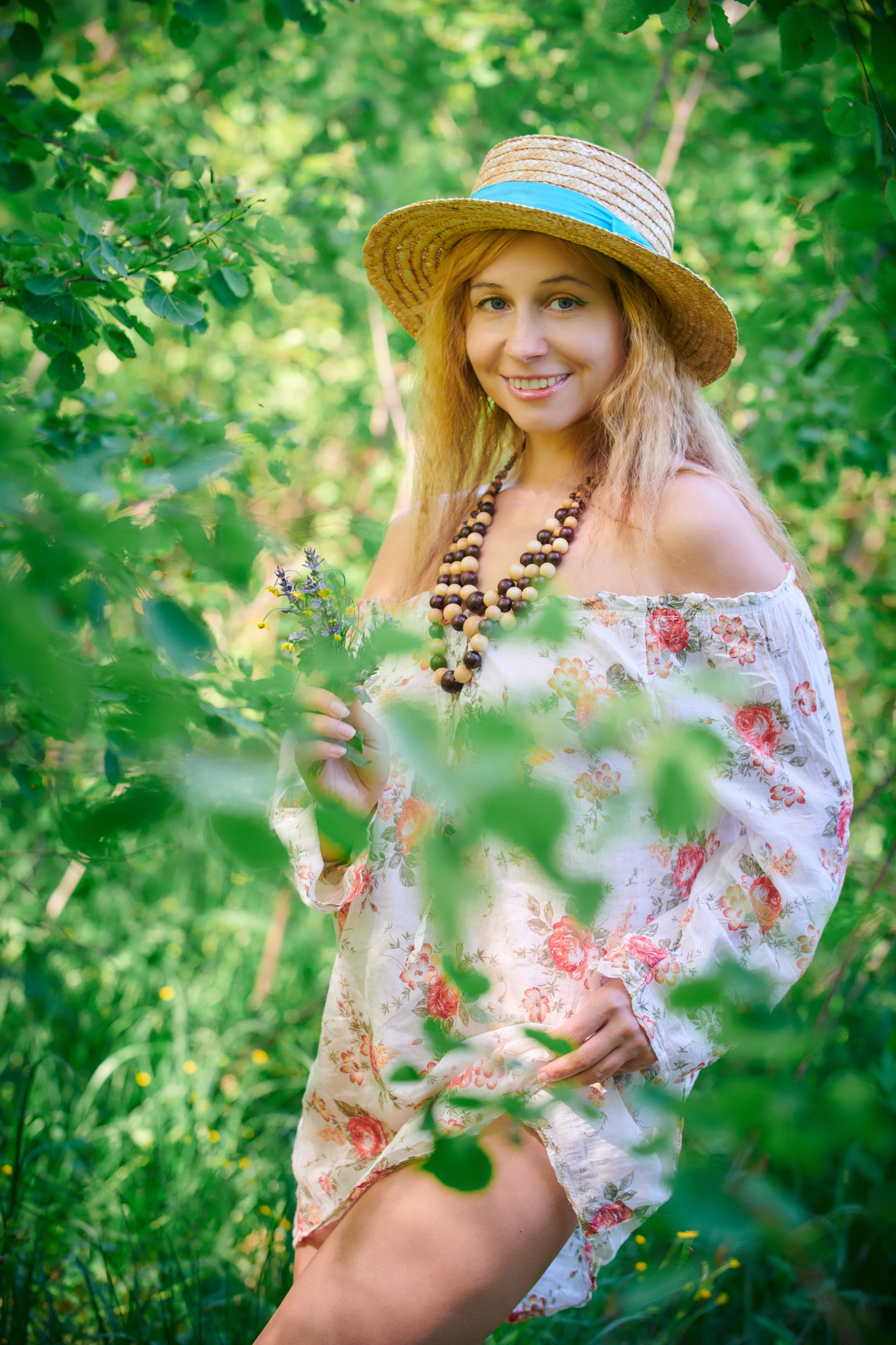Smiling woman in floral dress and hat enjoys nature in a lush summer setting.