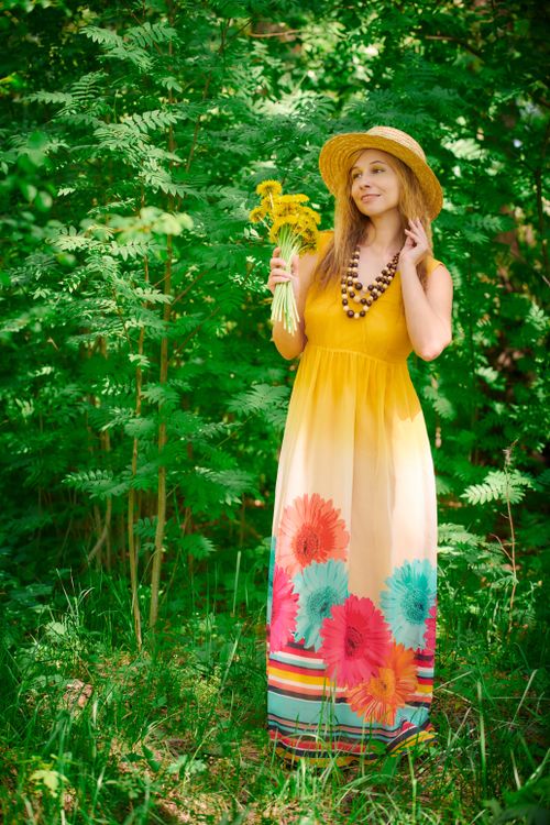 Free Woman wearing a straw hat and floral dress holding wildflowers in a lush green outdoor setting. Stock Photo