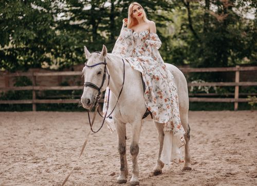 Free A woman in a floral dress riding a white horse in a sandy outdoor arena surrounded by nature. Stock Photo