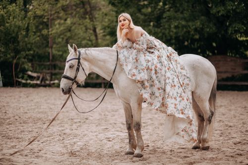 Free A woman in a floral dress elegantly poses on a white horse in an outdoor setting. Stock Photo