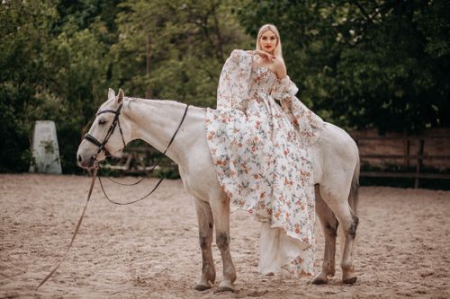 Free Stylish woman in floral dress elegantly seated on a white horse in an outdoor arena. Stock Photo