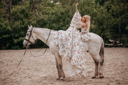 Free A stylish woman in a floral dress sits elegantly on a white horse in an outdoor setting. Stock Photo