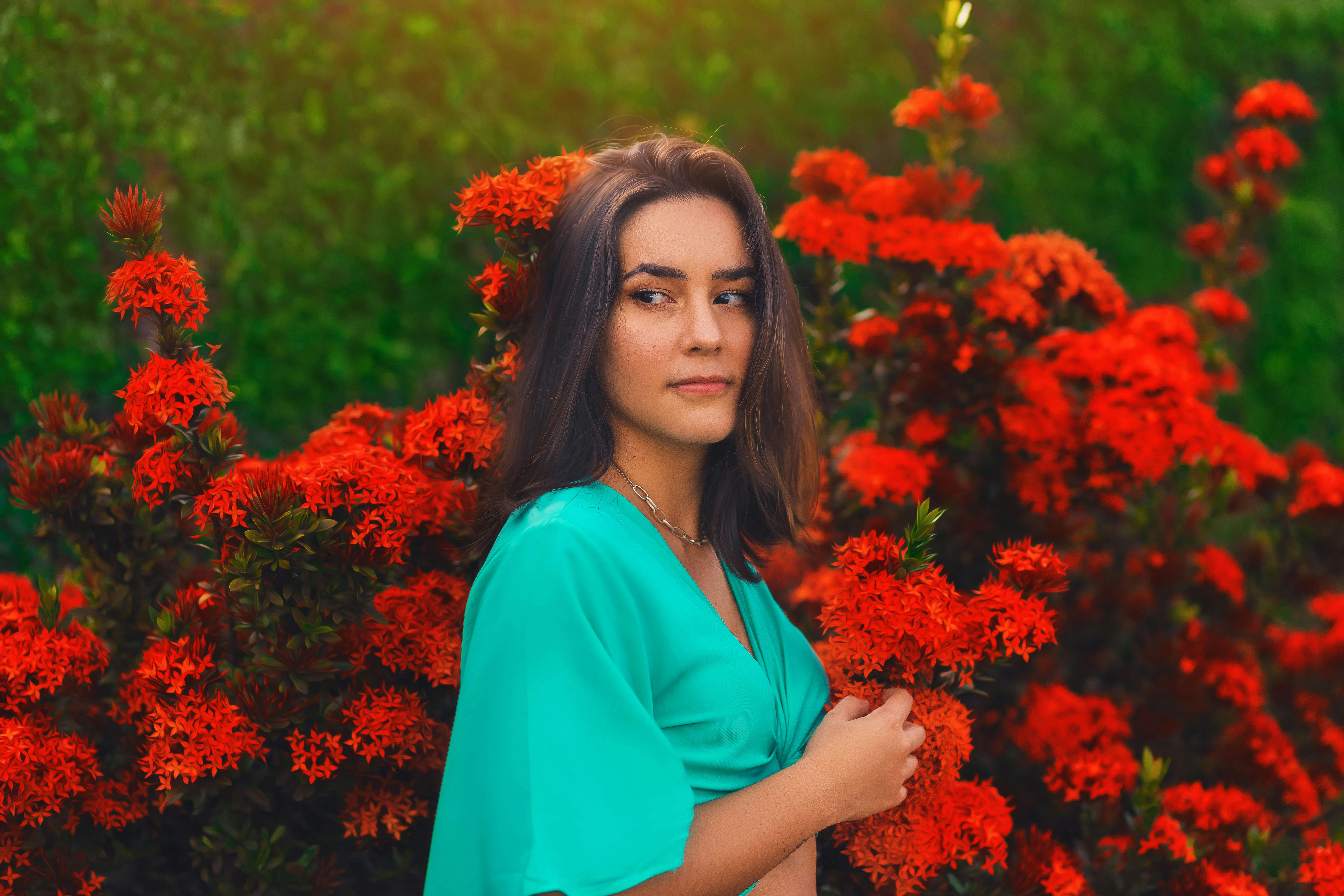 Woman in a teal dress posing amidst vibrant red flowers in a garden, Bauru, Brazil.