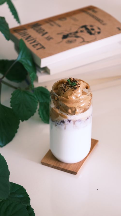 Free A delicious Dalgona coffee on a wooden coaster amidst books and greenery indoors. Stock Photo
