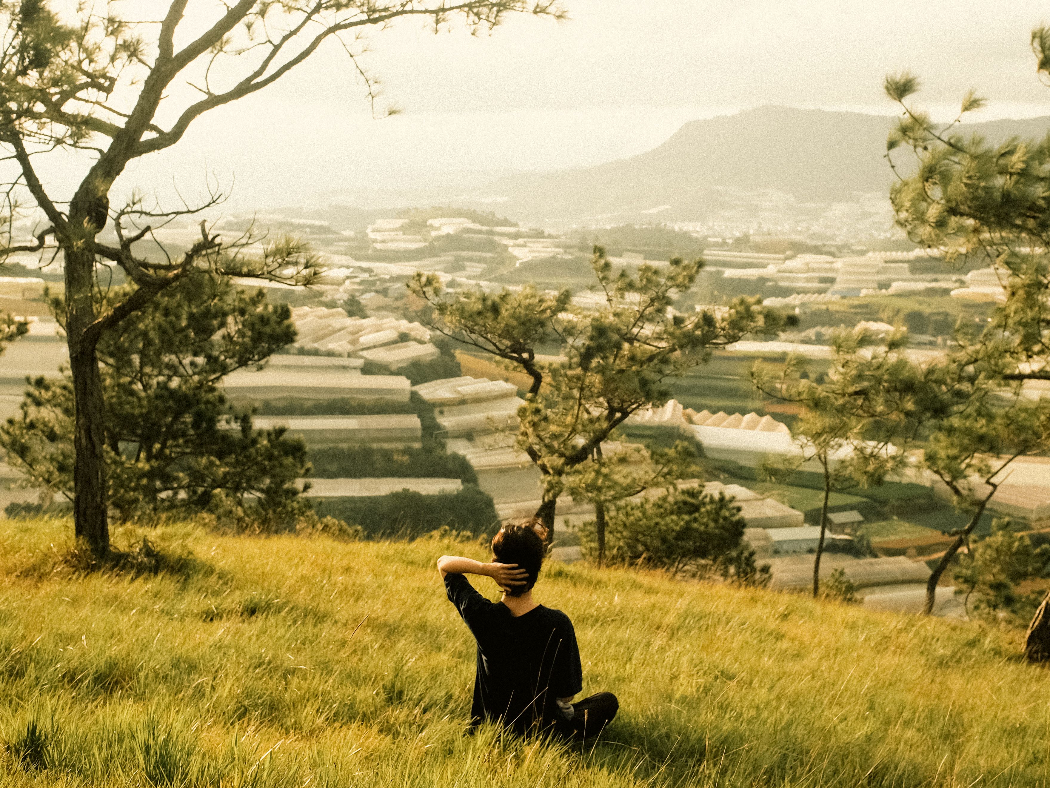 A woman sits in a grassy field overlooking a beautiful countryside landscape during sunset, embracing tranquility.