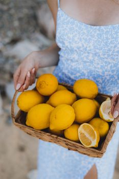 Woman in summer dress holding a basket of fresh yellow lemons outdoors, embodying rustic and tropical vibes.
