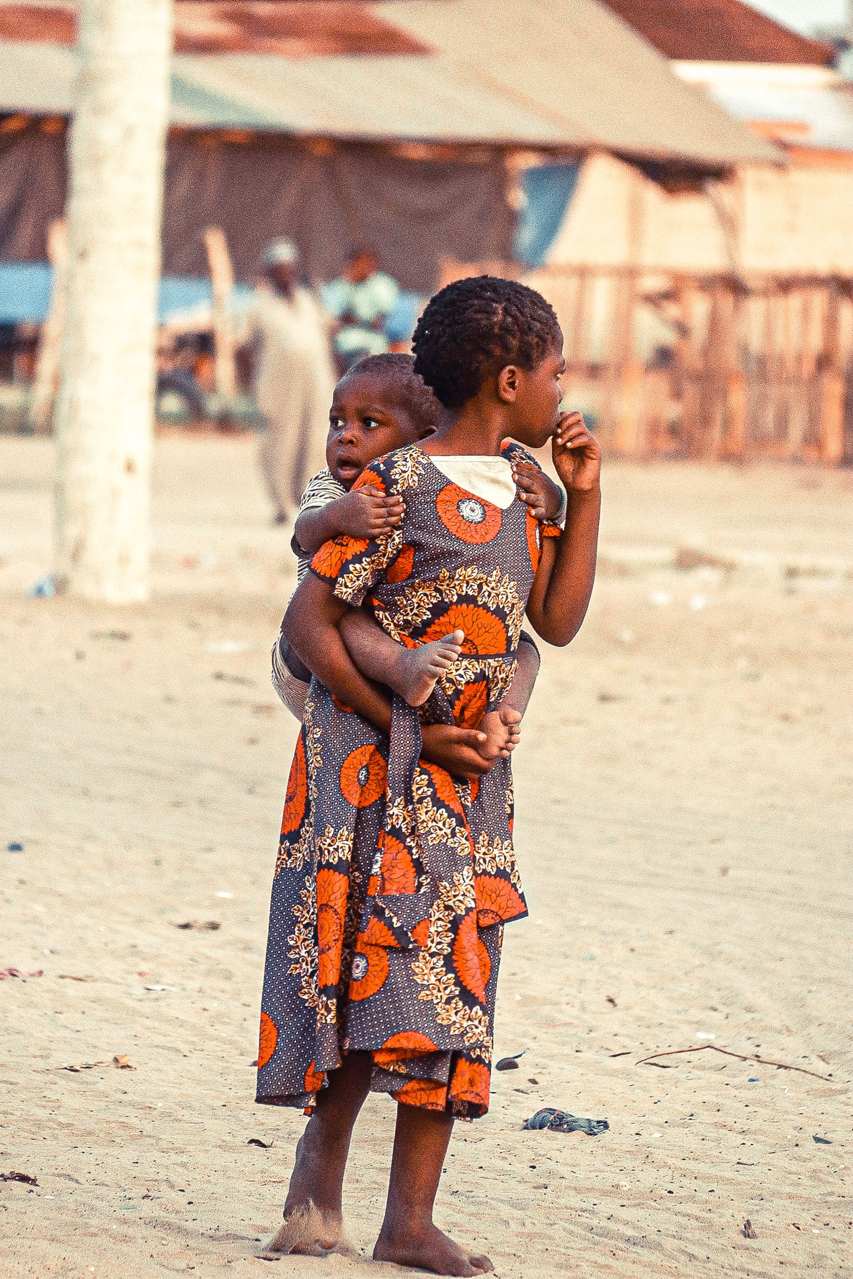 Young girl in traditional dress carries her sibling in an African village setting.