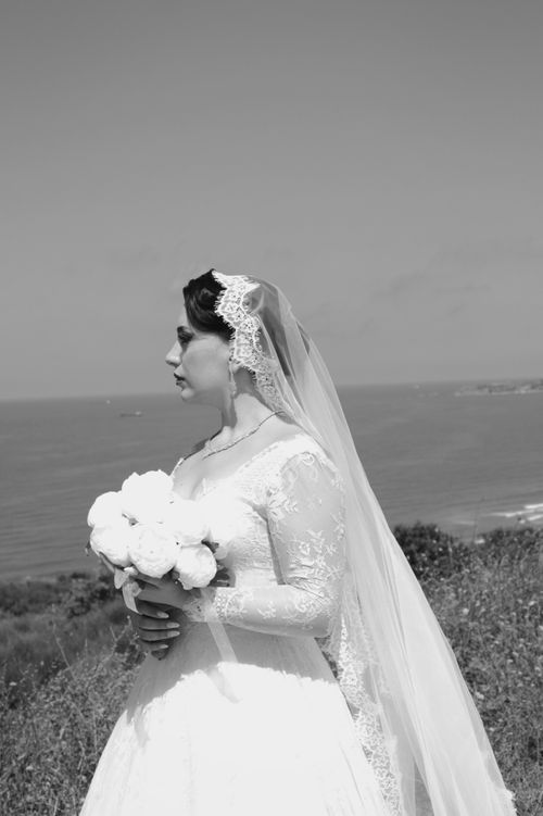 Free A beautiful bride in a lace dress holding flowers with an ocean backdrop. Stock Photo