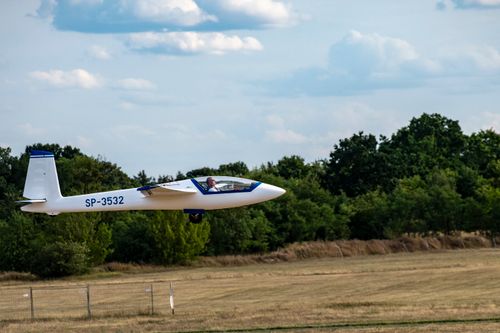 Free A glider plane in flight, soaring over a grassy airfield with trees in the background. Stock Photo