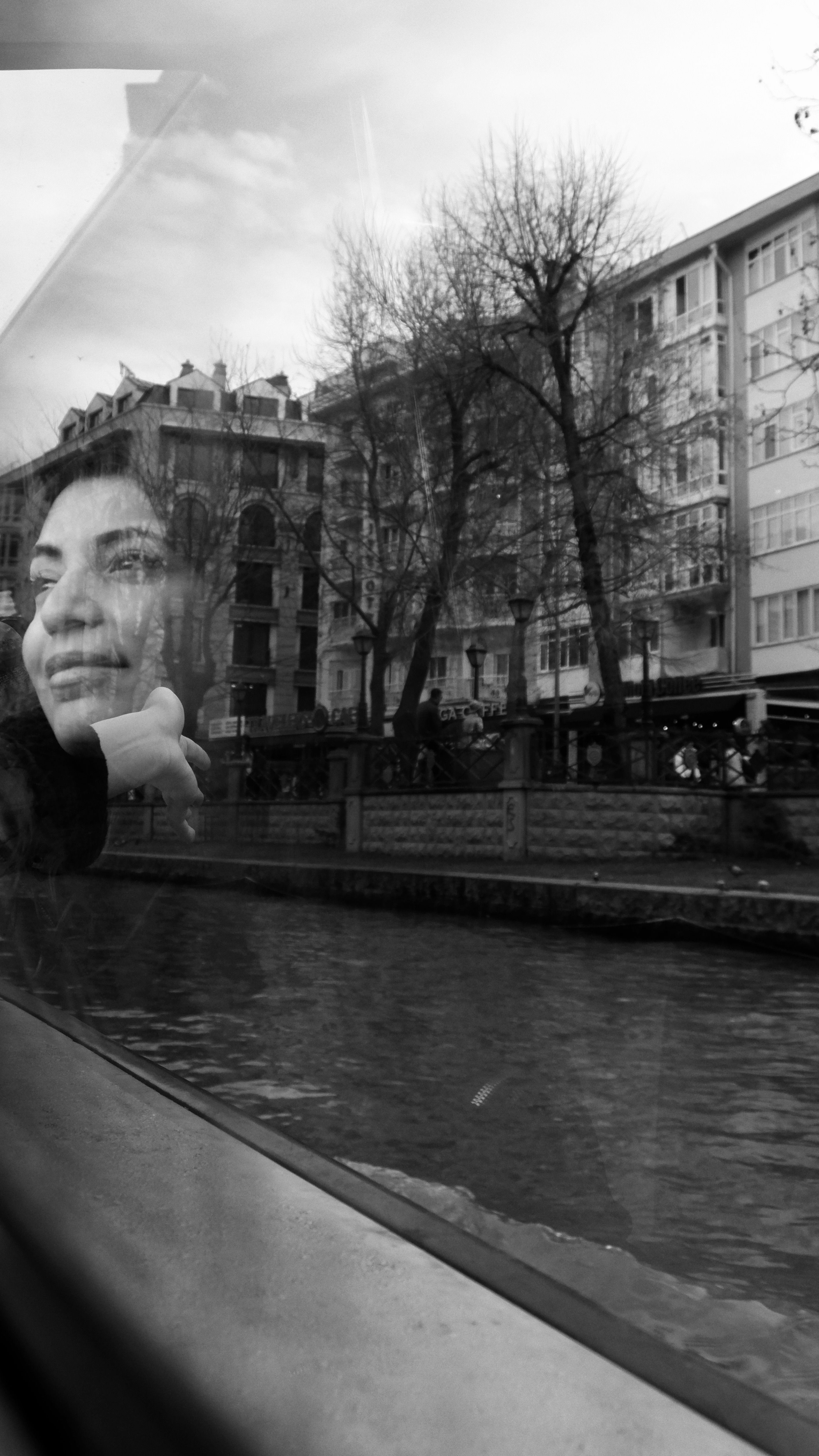 Black and white portrait of a woman reflected on a ferry window next to a city canal.